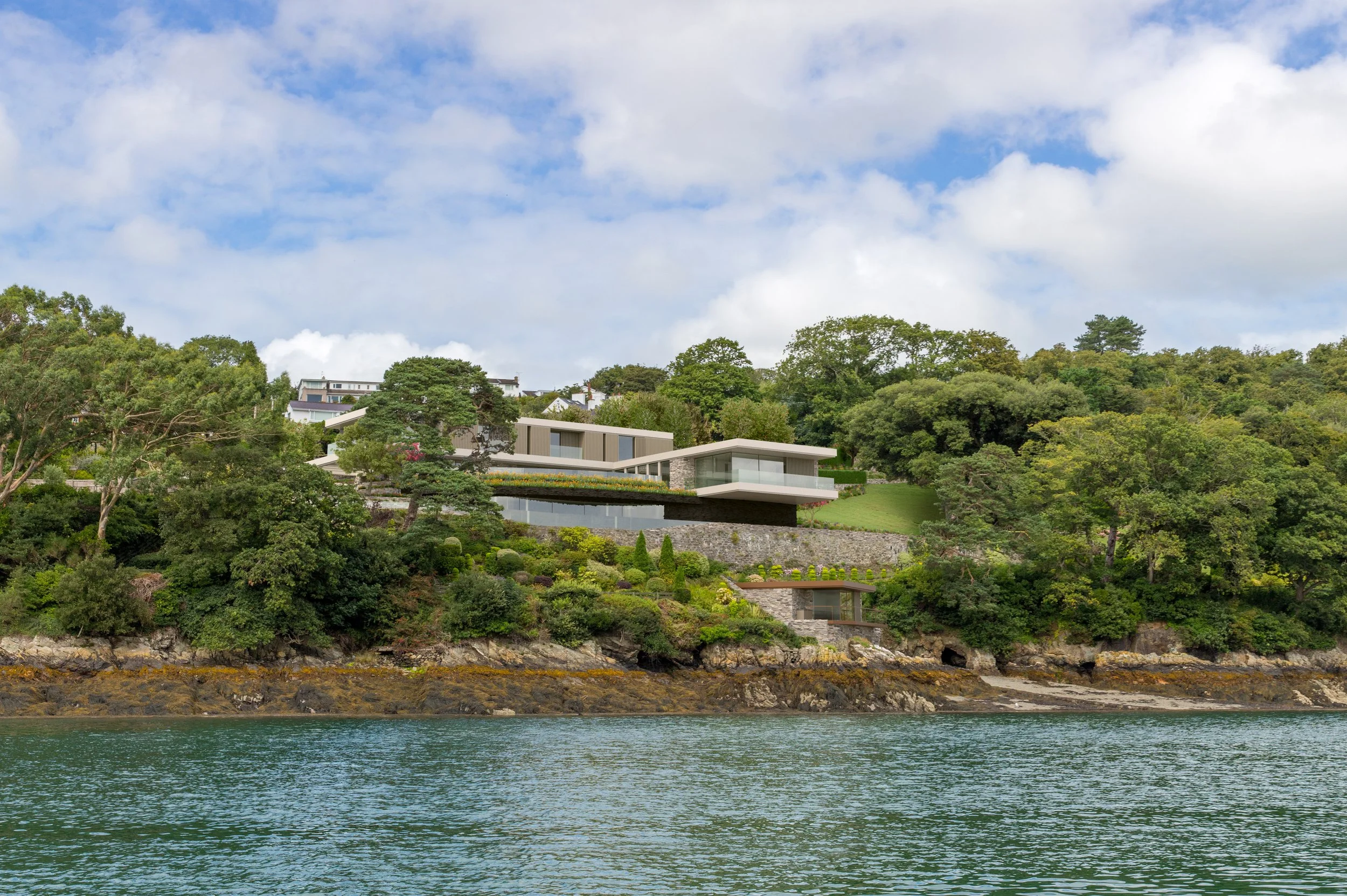 Modern house built on a hillside overlooking water, surrounded by lush trees and greenery, with a partly cloudy sky