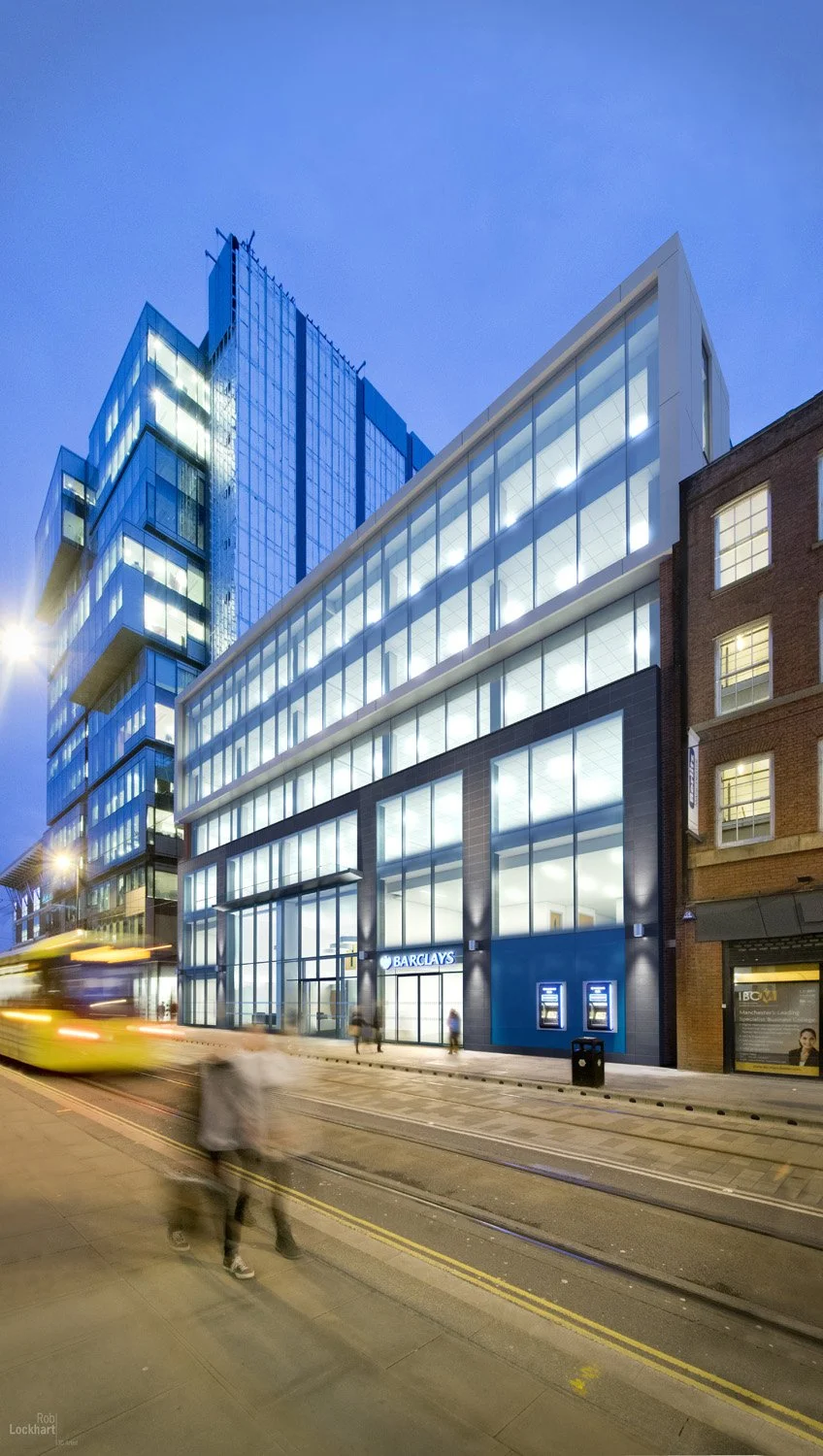 Modern glass office building with Barclays bank entrance on city street at dusk, blurred pedestrians and a yellow tram passing by.