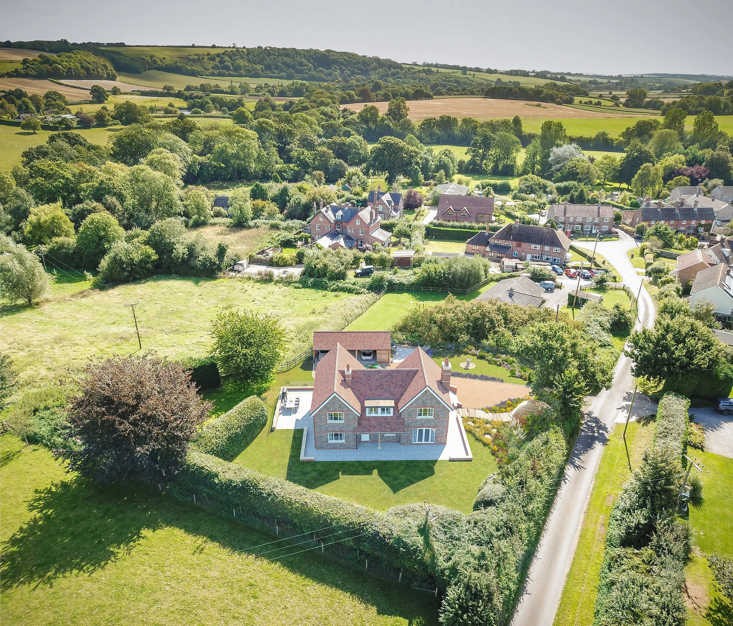Aerial view of a large brick house with a red tile roof, surrounded by a well-maintained lawn and garden, in a rural neighborhood with other houses, trees, and open fields.