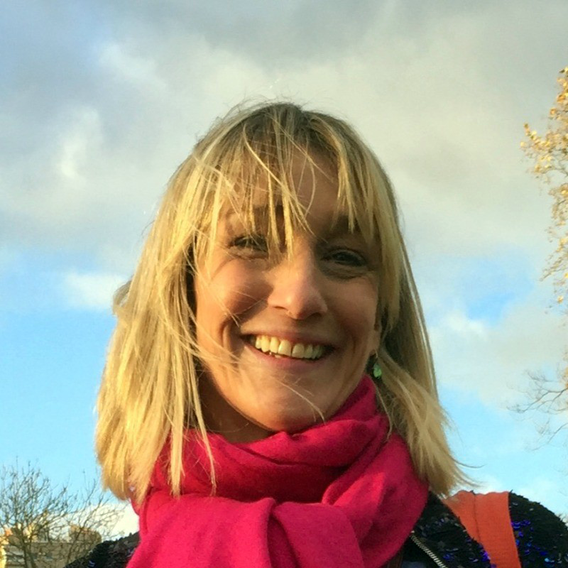 A smiling woman with blonde hair, wearing a bright pink scarf, standing outdoors with a blue sky and some trees in the background.