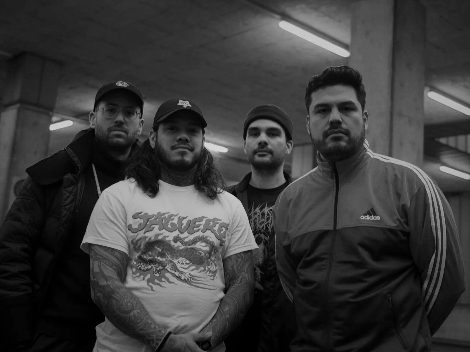 Vier junge Männer stehen in einer dunklen, industriellen Umgebung, alle schauen ernst in die Kamera.