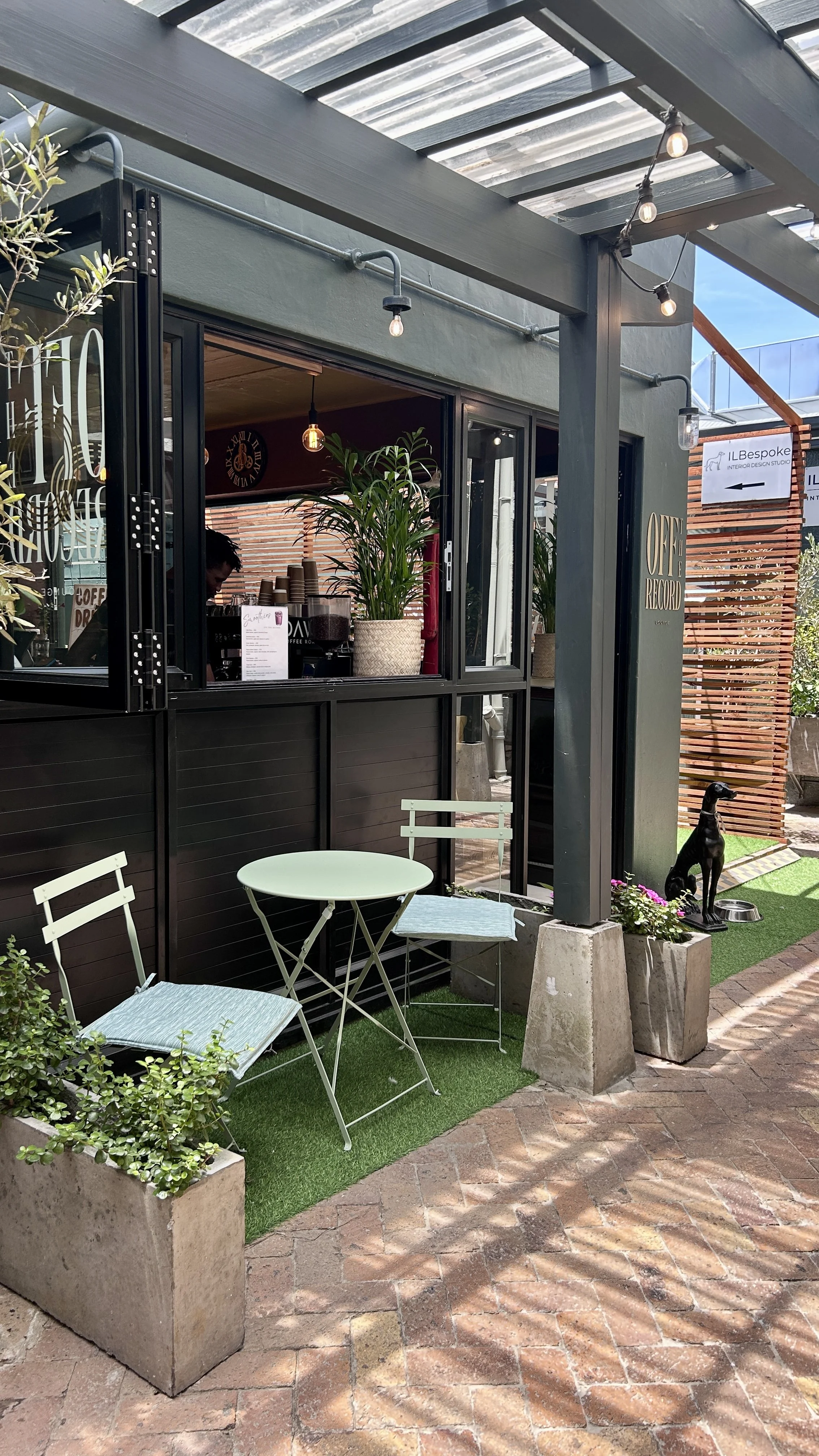 Outdoor cafe seating with two light blue chairs and a small round table, a potted plant, and a dog statue, next to a coffee shop with a black exterior, open window, and string lights.