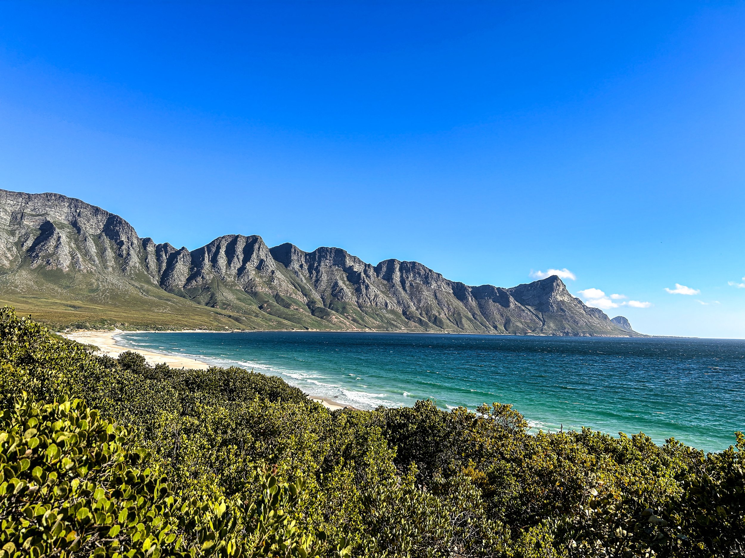 A scenic view of a rocky mountain range along a coastline with a sandy beach and green shrubbery, under a clear blue sky.