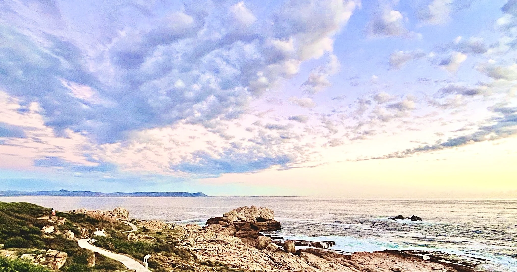 Scenic coastal view with rocky shoreline, calm water, and a partly cloudy sky during sunset.