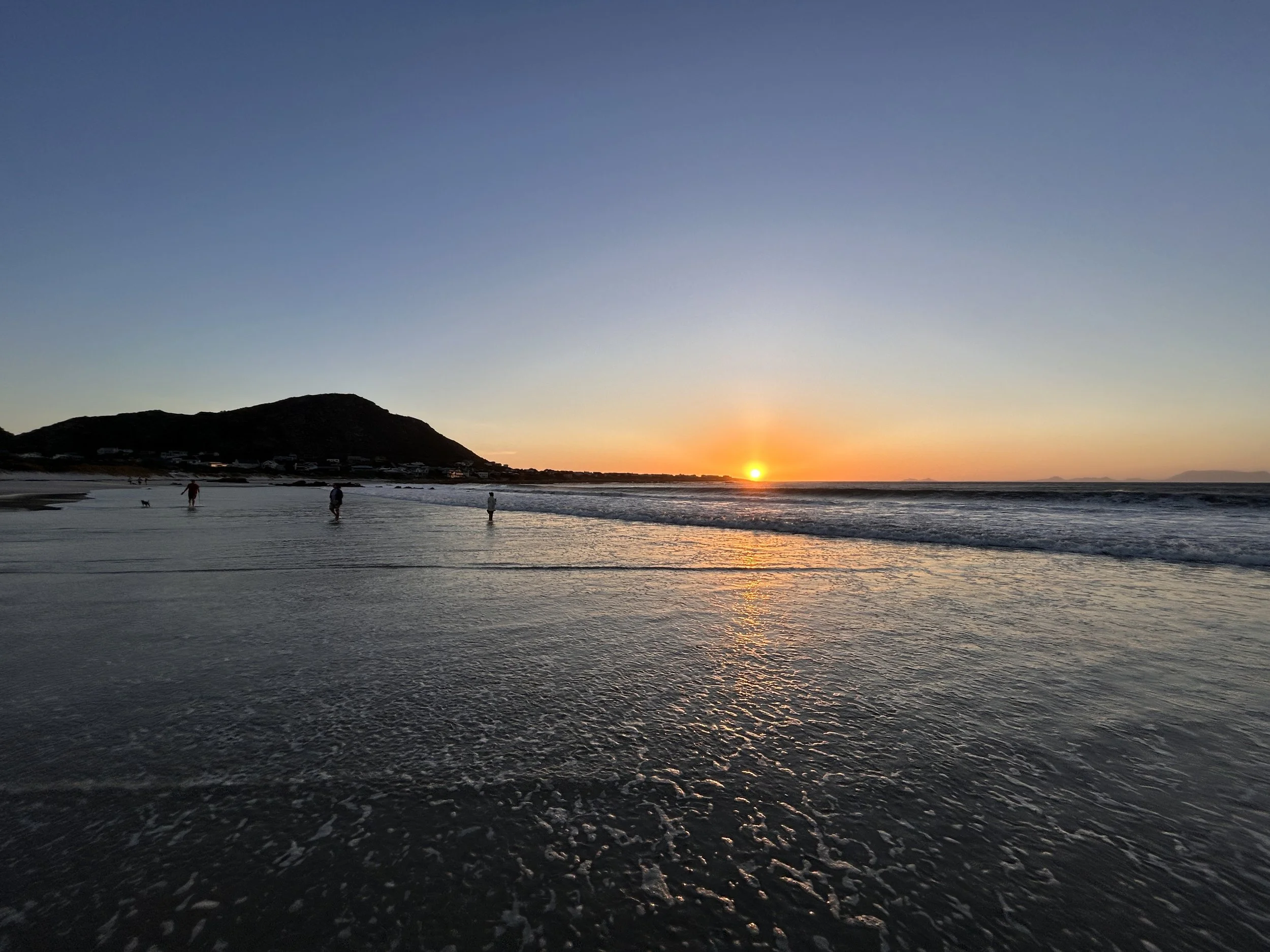 Sunset at the beach with few people walking along the shoreline, calm waves, and a hill in the background.