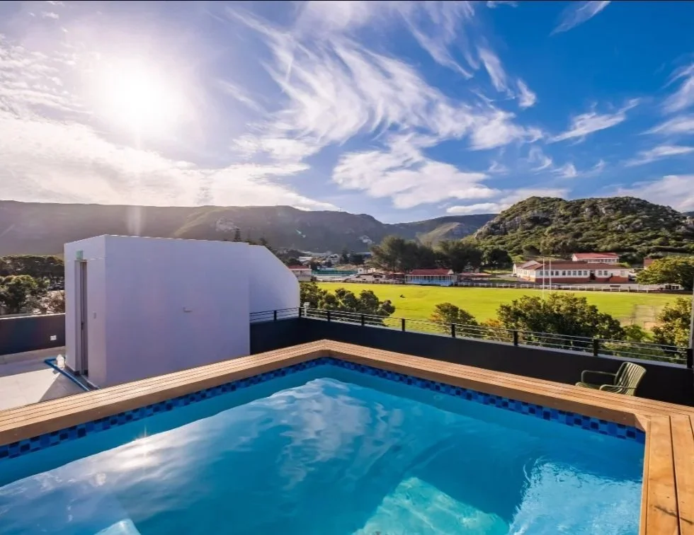A rooftop pool with a wood deck, overlooking a green field with trees and buildings, under a partly cloudy sky with the sun shining.