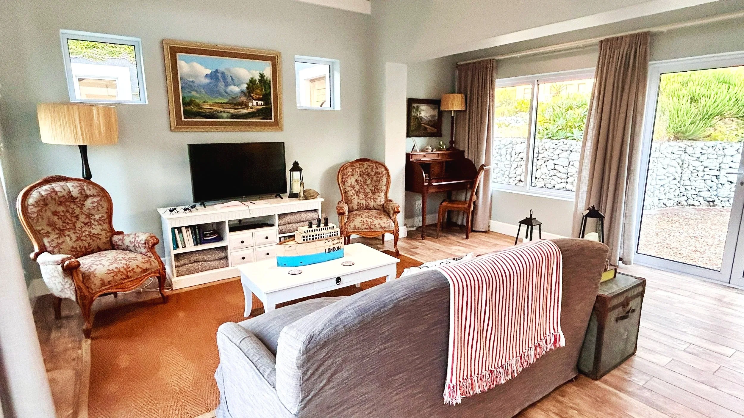 Living room with sofa, armchairs, coffee table, TV, vintage desk and chair, large window, and sliding glass door leading outside