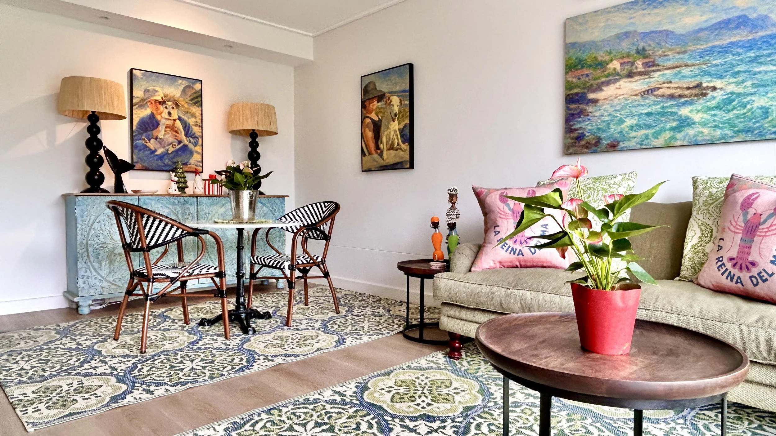 A cozy living room with a beige sofa holding decorative pink pillows featuring a lobster illustration. A round wooden coffee table with a potted plant sits in front of the sofa. Artwork adorns the walls, including beach scenes and a woman with a dog. A blue sideboard with lamps, a sculpture, and flowers is against the wall. A patterned area rug covers part of the flooring, and a small side table holds decorative figurines.