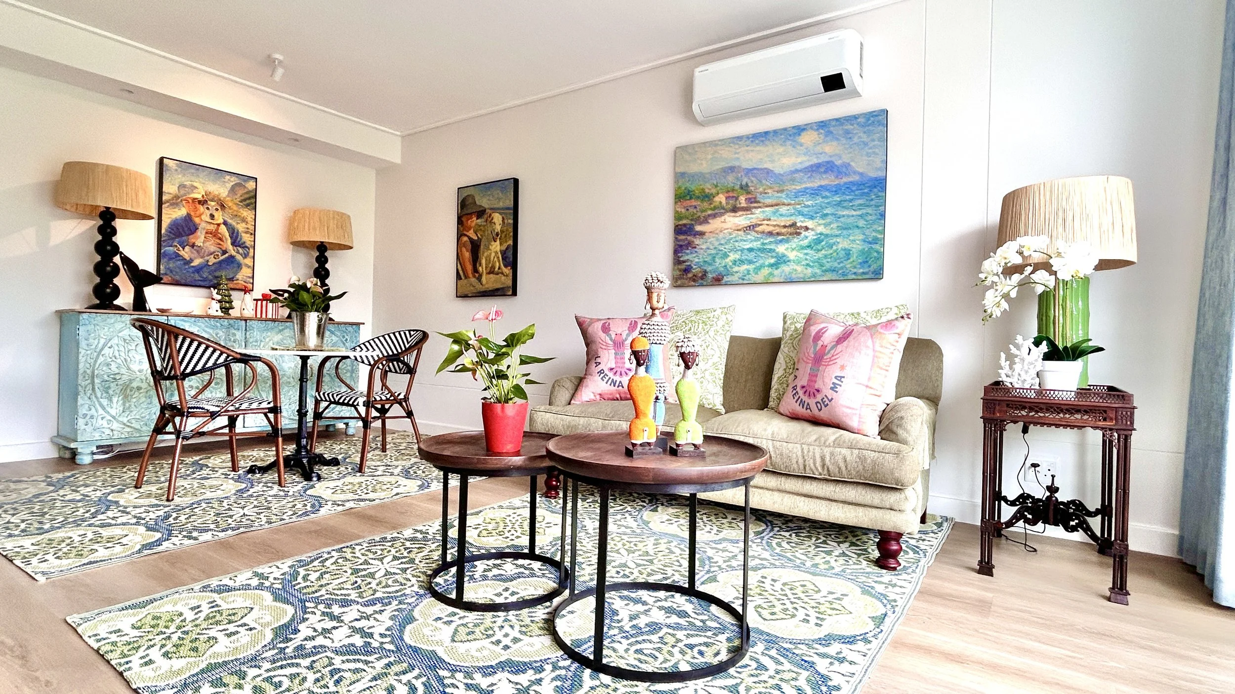 A living room with a beige sofa, colorful throw pillows, and a round wooden coffee table with decorative objects. There are paintings on the wall, a blue sideboard with lamps, and a potted plant. The room has a patterned rug and wooden flooring.