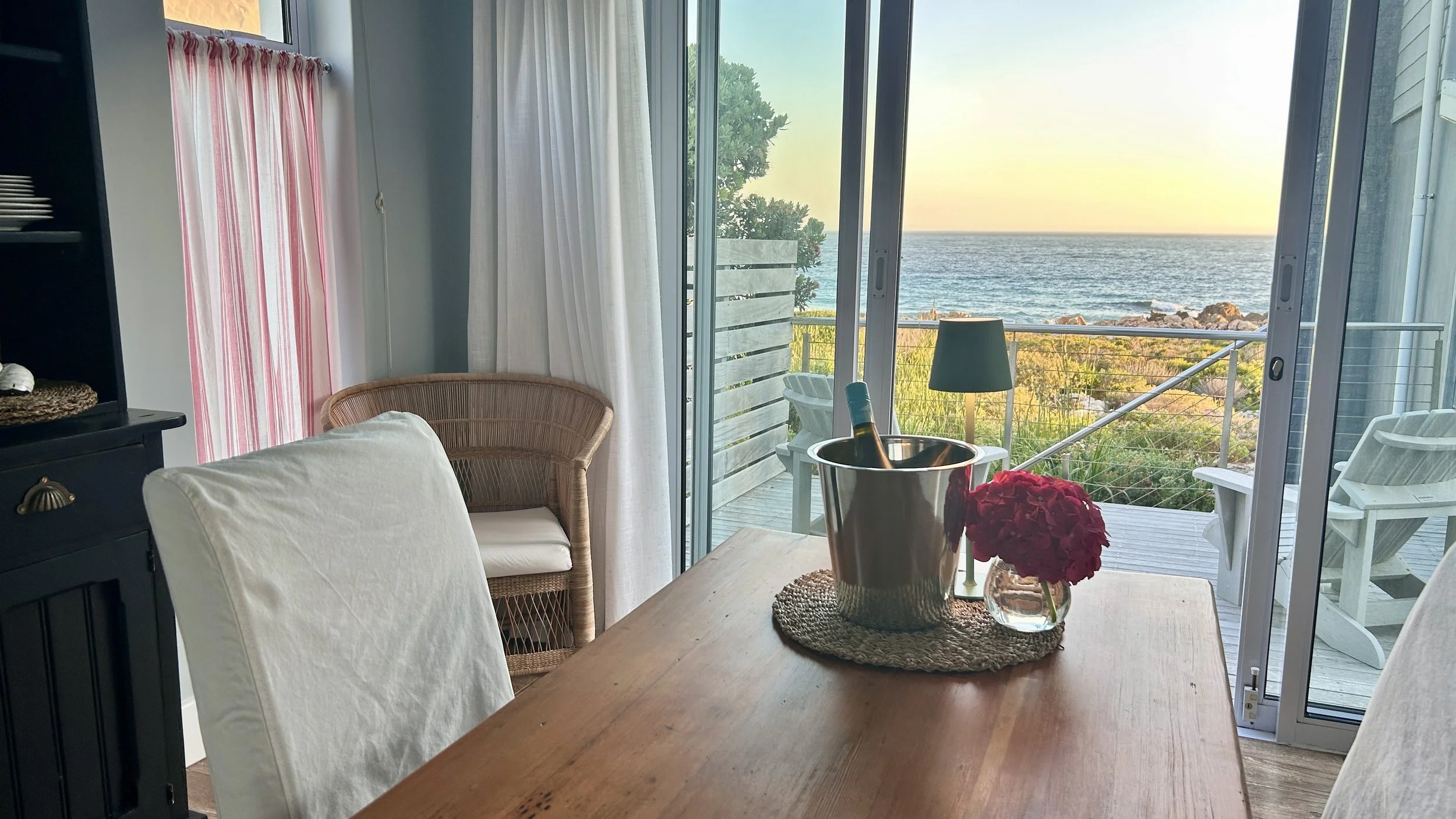 A dining room with a wooden table, white chairs, and a view of the ocean through sliding glass doors. The table has a metal ice bucket with a bottle of wine and a small vase with pink flowers. Outside, there is a small deck with white Adirondack chai
