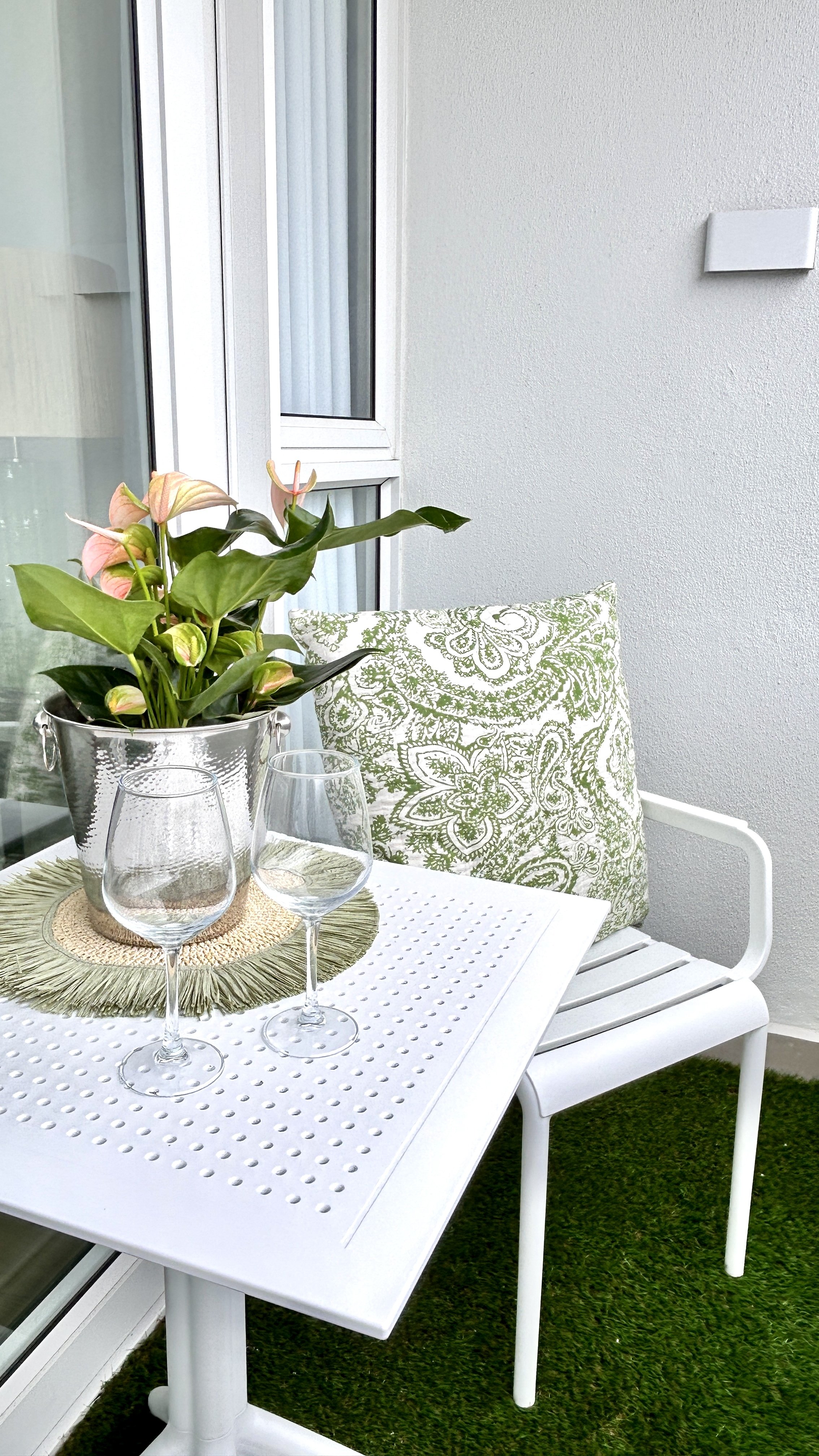 A white outdoor patio table with two empty wine glasses, a potted pink and green flowering plant, and a green and white patterned pillow on a white chair. The table is on green grass, next to a white wall and sliding glass door.