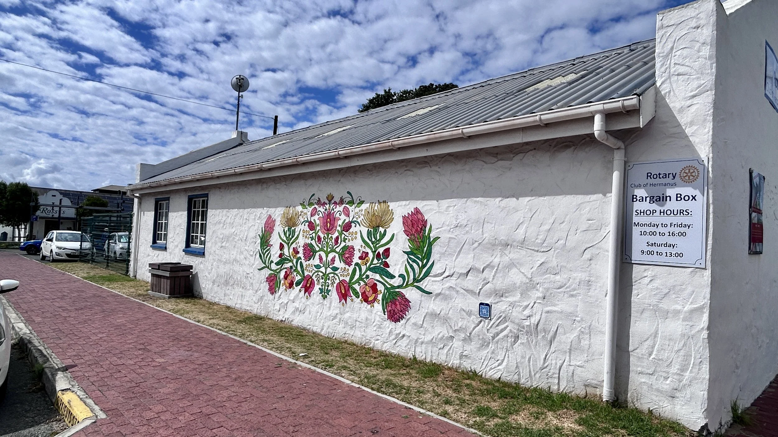 White building with a colorful floral mural on the wall, a sidewalk with parked cars nearby, a sign indicating Rotary Club Hermanu's bargain box and shop hours, under a partly cloudy sky.