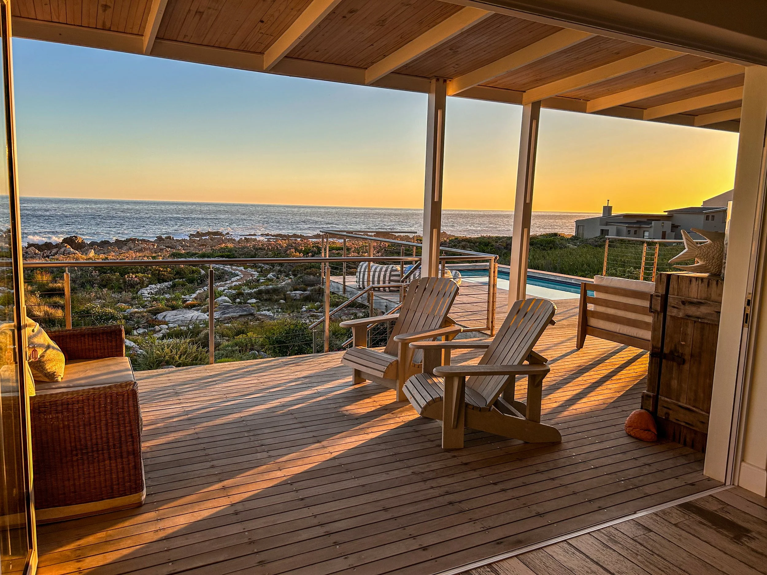 Ocean view from a wooden deck with Adirondack chairs, a wicker chair, and a small wooden cabinet at sunset.