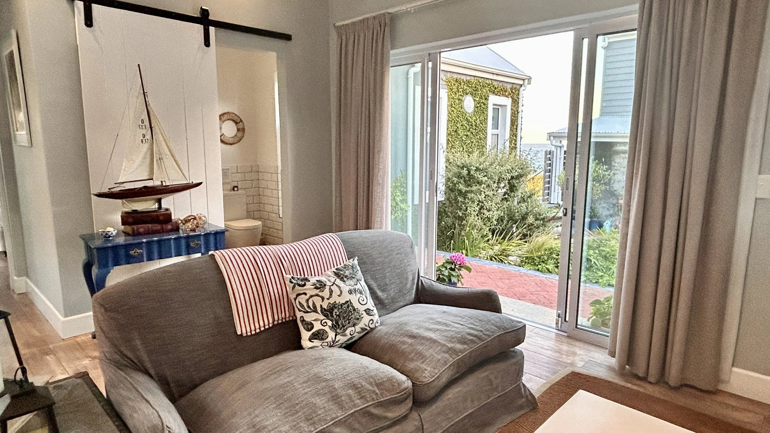 Living room with large sliding glass doors leading to an outdoor stone patio, beige curtains, striped and floral cushions on a gray sofa, a small coffee table with lanterns and books, a blue chest with a model sailboat and books, and a red cabinet wi