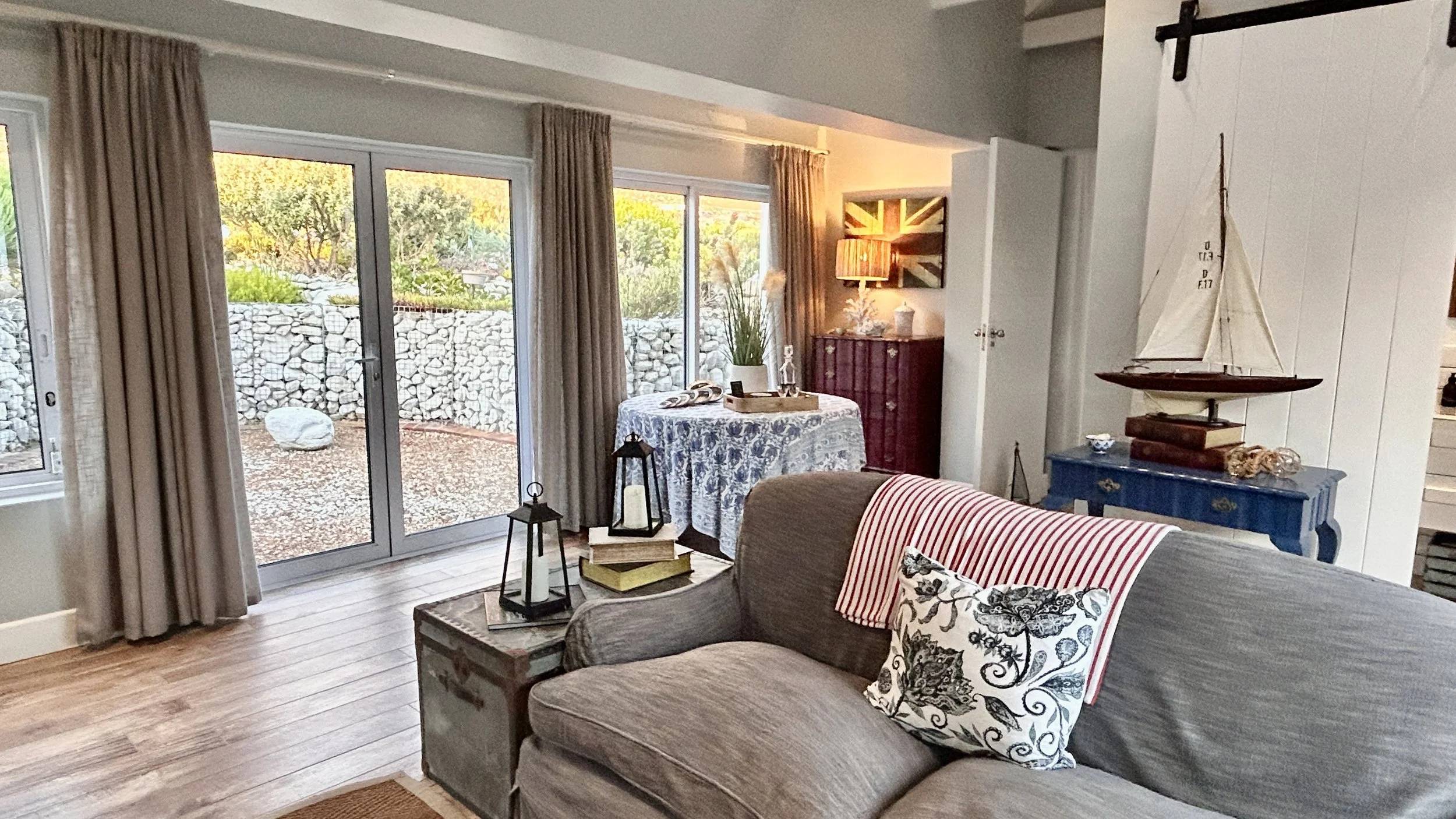 Living room with sliding glass door leading outside, beige curtains, a sofa with striped and floral pillows, a round table with a lace tablecloth, decorative lanterns, and a blue side table with a model sailboat and books.