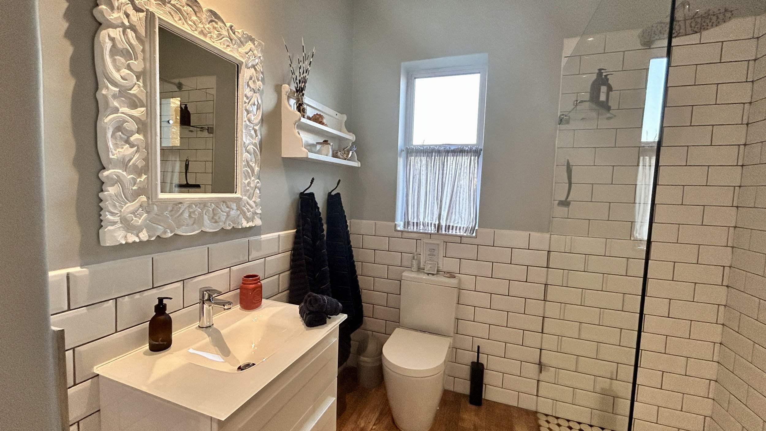 A modern bathroom with white subway tiles on the walls, a white vanity with a sink, a large ornate white mirror, a window with a sheer curtain, two black towels hanging, and a glass shower enclosure with white tiles and a shelf with toiletries.