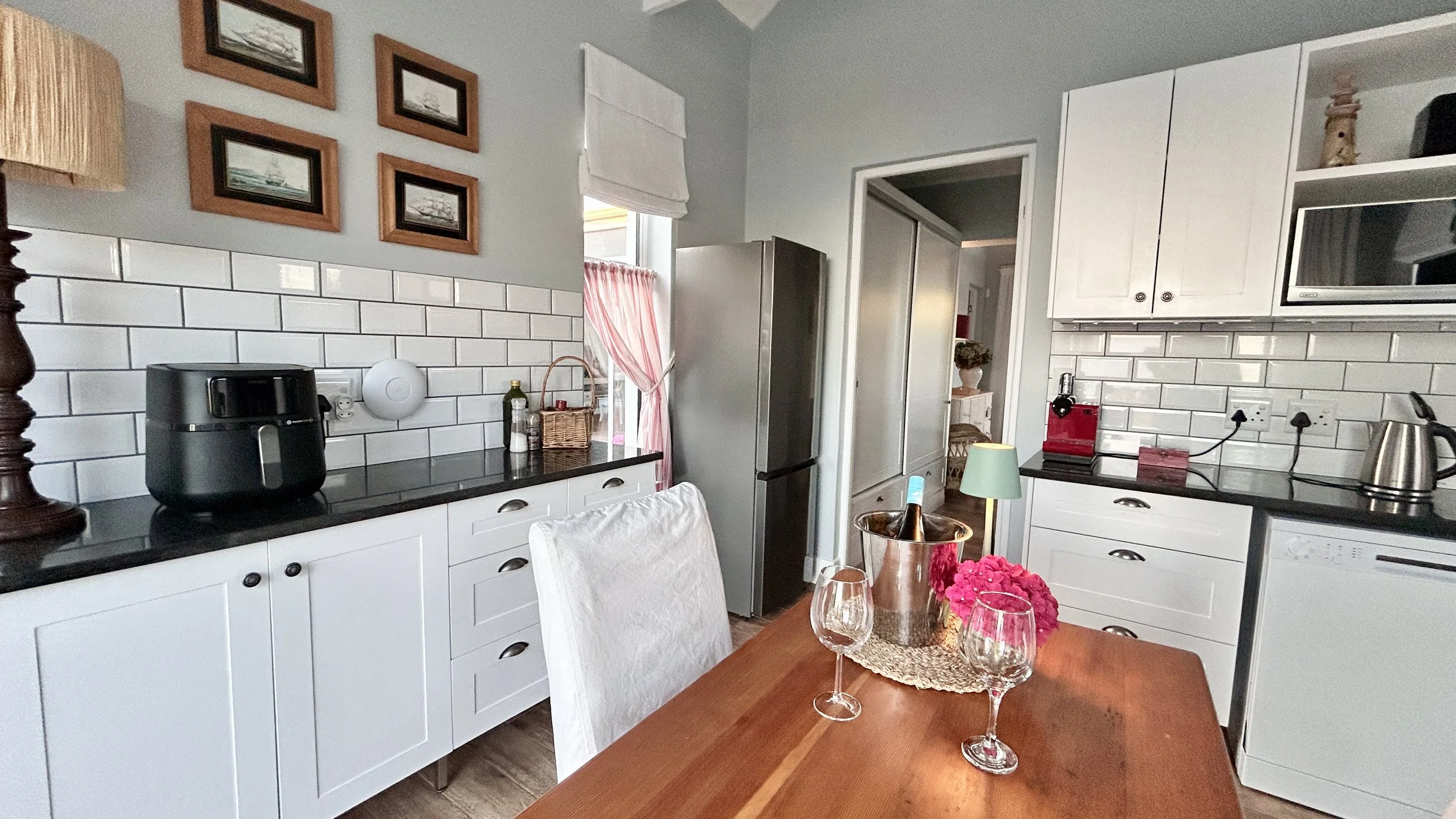 Kitchen with white cabinets, black countertops, and a white subway tile backsplash. A dining table with a pink flower centerpiece, two wine glasses, and a bucket of wine in the foreground. Appliances include a refrigerator, microwave, air fryer, toas