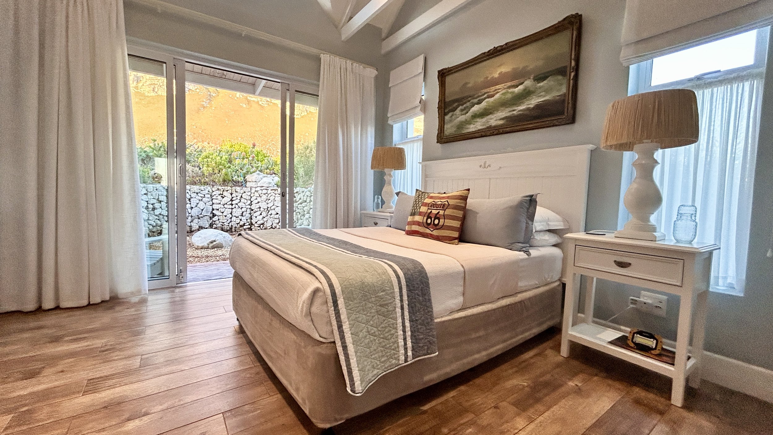 A cozy bedroom with a white bed, two nightstands with lamps, a Route 66 pillow, nature outside sliding glass door, and a framed painting of ocean waves.