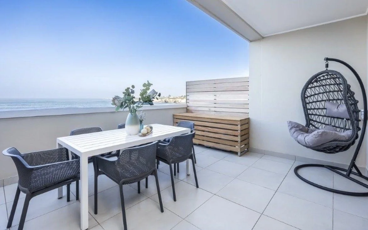 Balcony with a white dining table, six black wicker chairs, a vase with greenery, a wooden bench, and a hanging cushioned egg chair overlooking the ocean under a clear blue sky.