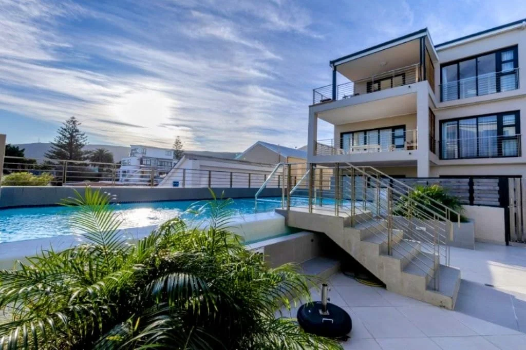 Modern apartment building with a rooftop pool, glass railings, and outdoor stairs leading to the pool area. There are green plants in the foreground and additional buildings and trees in the background under a partly cloudy sky.