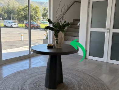 Interior of a modern building with a round black table, decorated with a large white vase holding green foliage and tall decorative branches, positioned on a circular gray rug near glass doors and windows showing an outdoor parking lot with a few cars and trees.