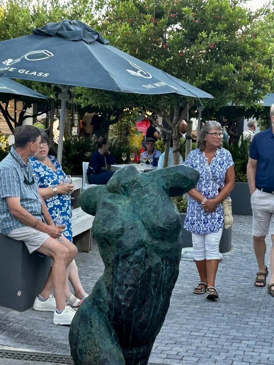 People socializing outdoors around a bronze dog sculpture with an umbrella and trees in the background.