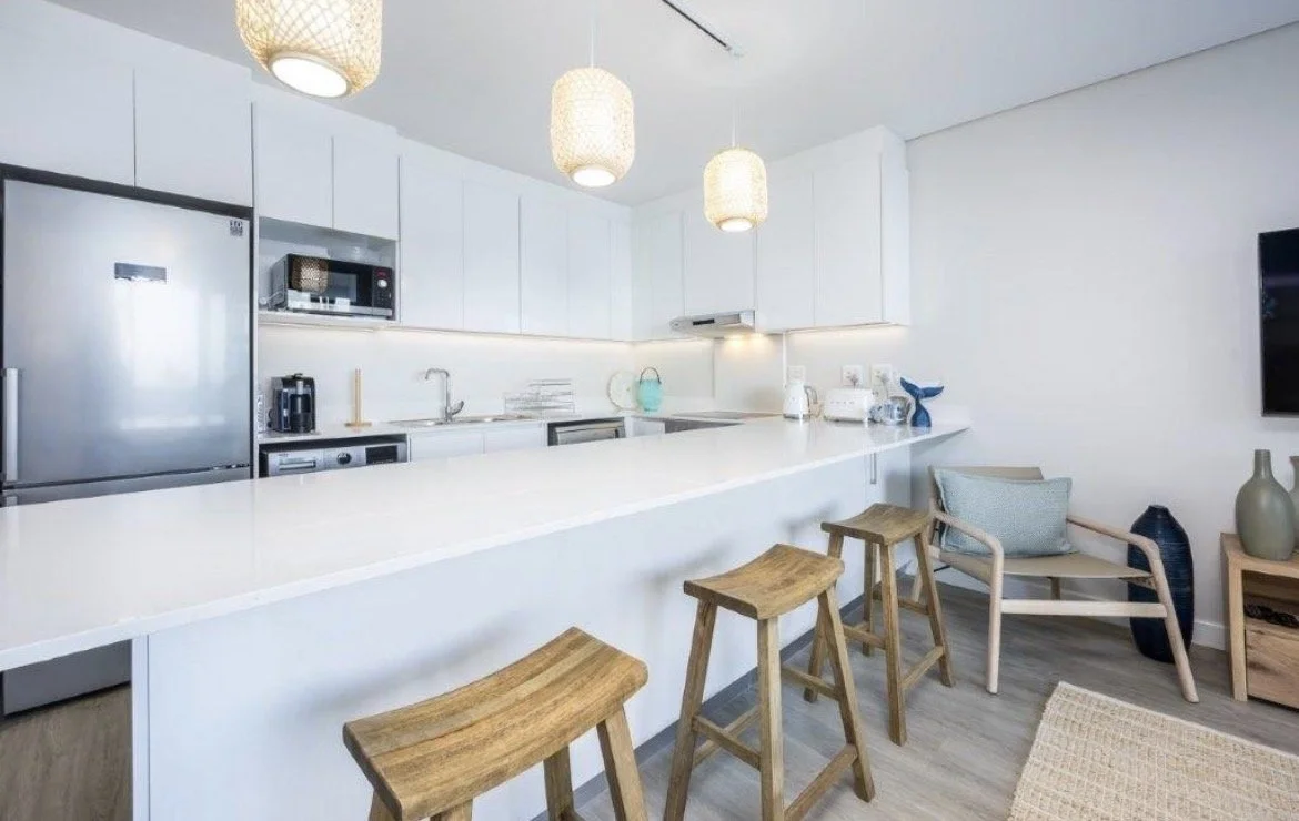 Modern kitchen with white cabinets, a stainless steel refrigerator, microwave, coffee maker, and a white countertop with bar stools and a seating area with a chair and large vases.