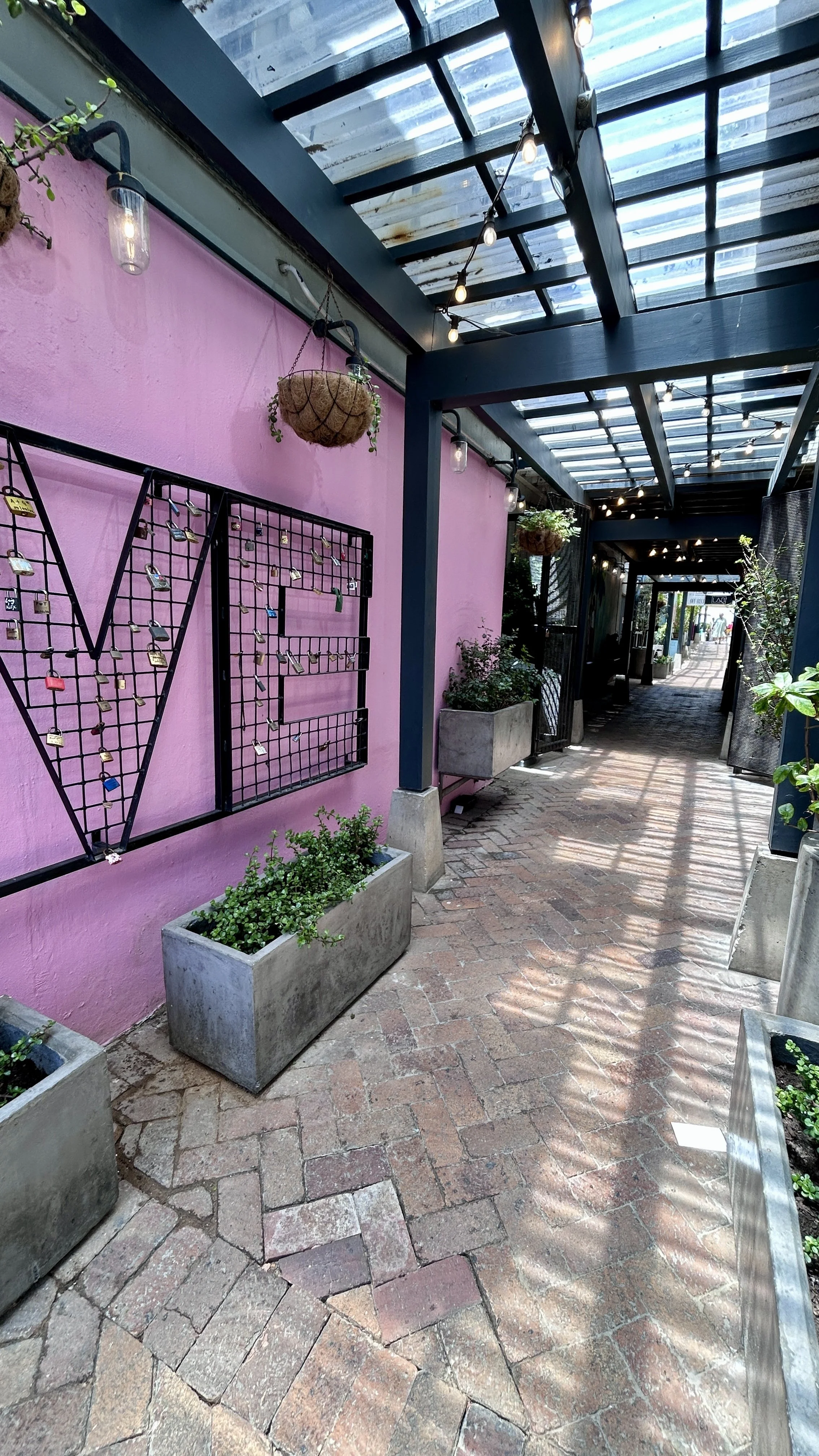 An outdoor walkway with a pink wall decorated with a metal frame installation with locks, potted plants, and hanging baskets. The walkway has brick paving and a glass roof supported by blue beams, with string lights hanging overhead.