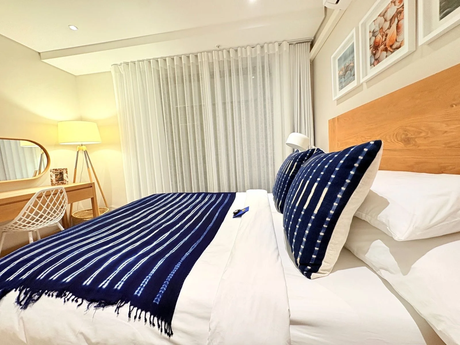 Hotel room with a neatly made bed, blue and white striped pillows and a matching throw, a wooden headboard, white curtains, and a desk with a lamp and decorative items.
