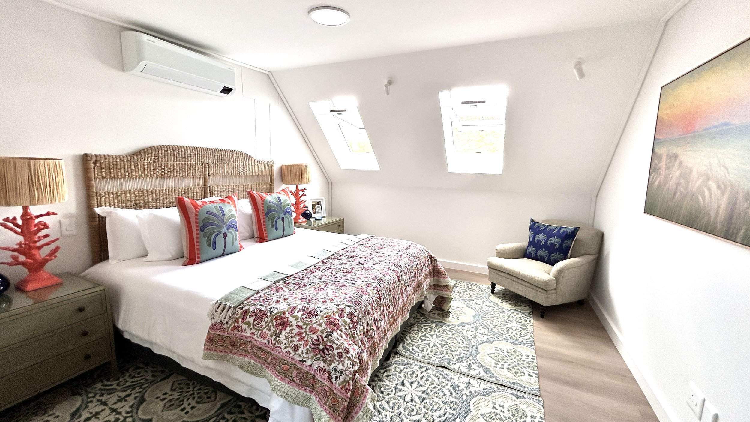 A bedroom with a bed featuring colorful pillows, a floral bedspread, and nightstands with lamps. There is a wicker headboard, a small armchair, and two skylight windows.