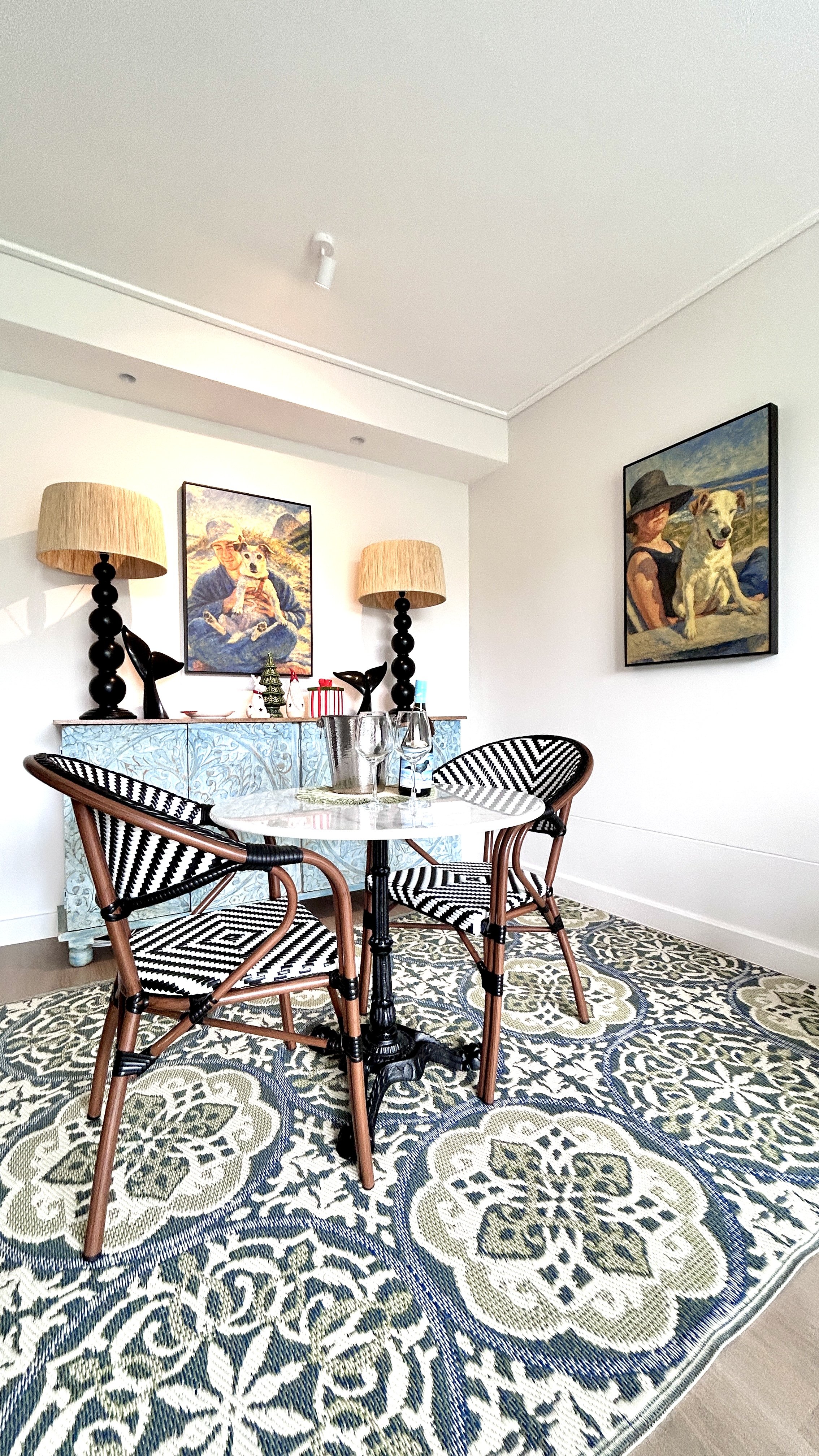 A dining area with a round glass table, three black and white striped chairs, a patterned rug, and a blue decorative sideboard. There are two paintings of women and dogs on the wall, with two matching lamps on the sideboard.
