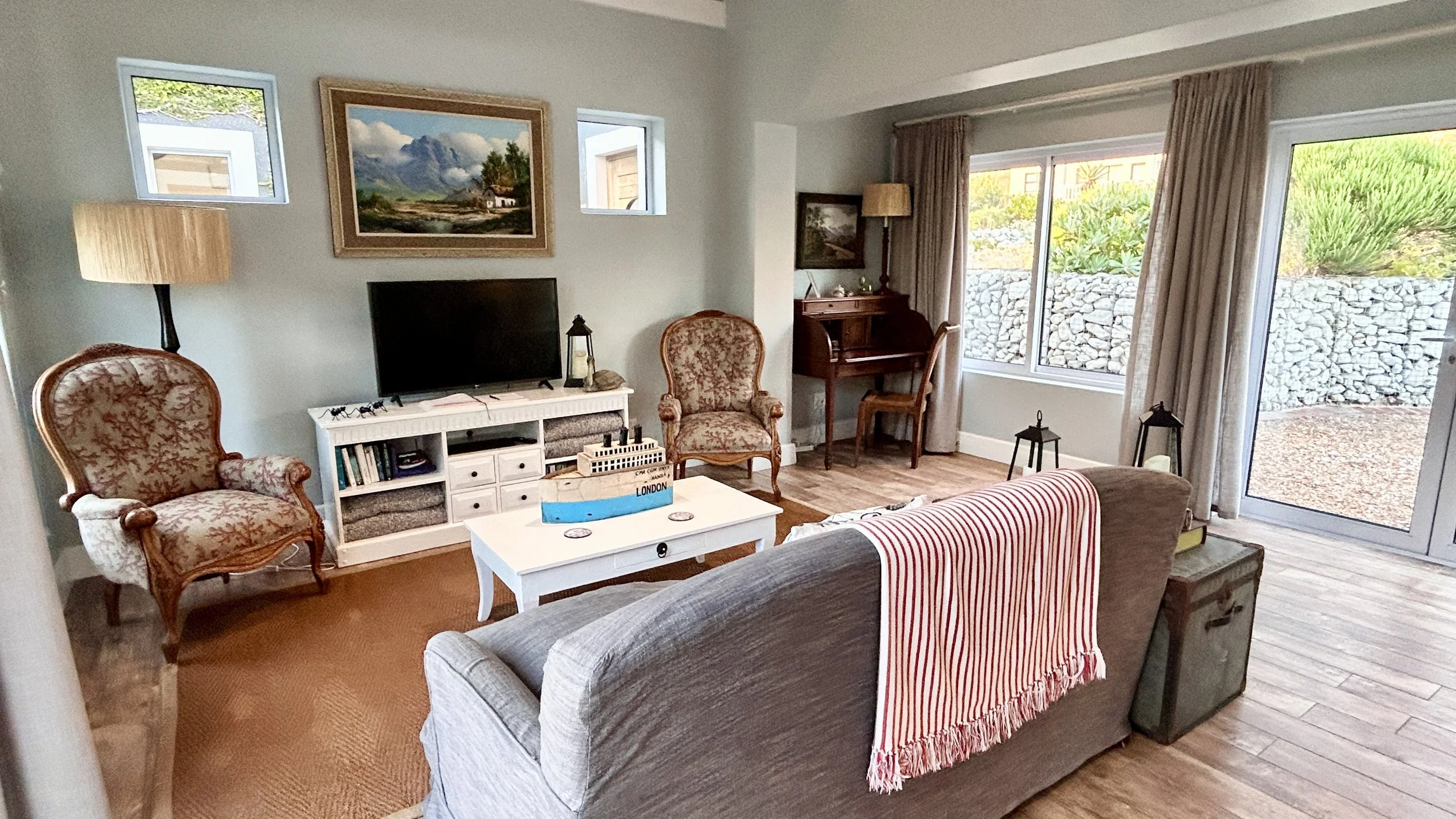 Living room with sofa, armchairs, coffee table, TV, vintage desk and chair, large window, and sliding glass door leading outside