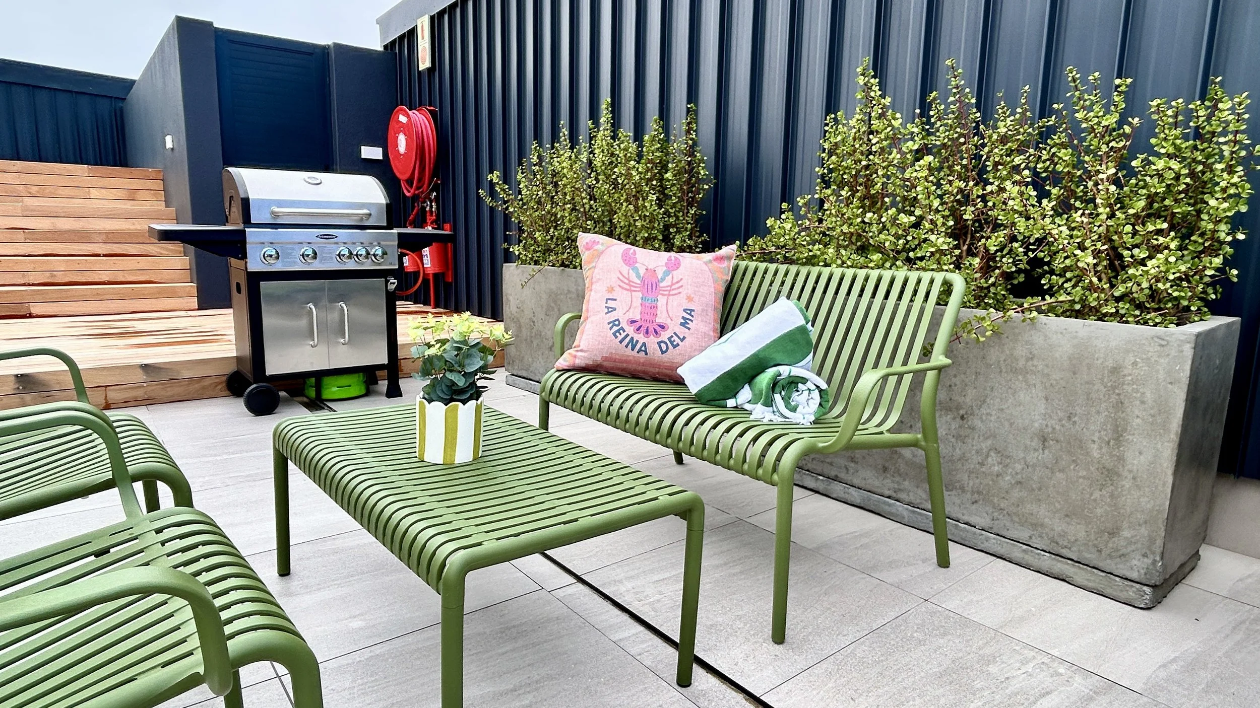 Outdoor patio area with green metal furniture, including a bench and two chairs, a wooden table, potted plant, large concrete planter with bushes, charcoal grill, red fire hose reel, and a wooden staircase, all enclosed with a dark blue fence.