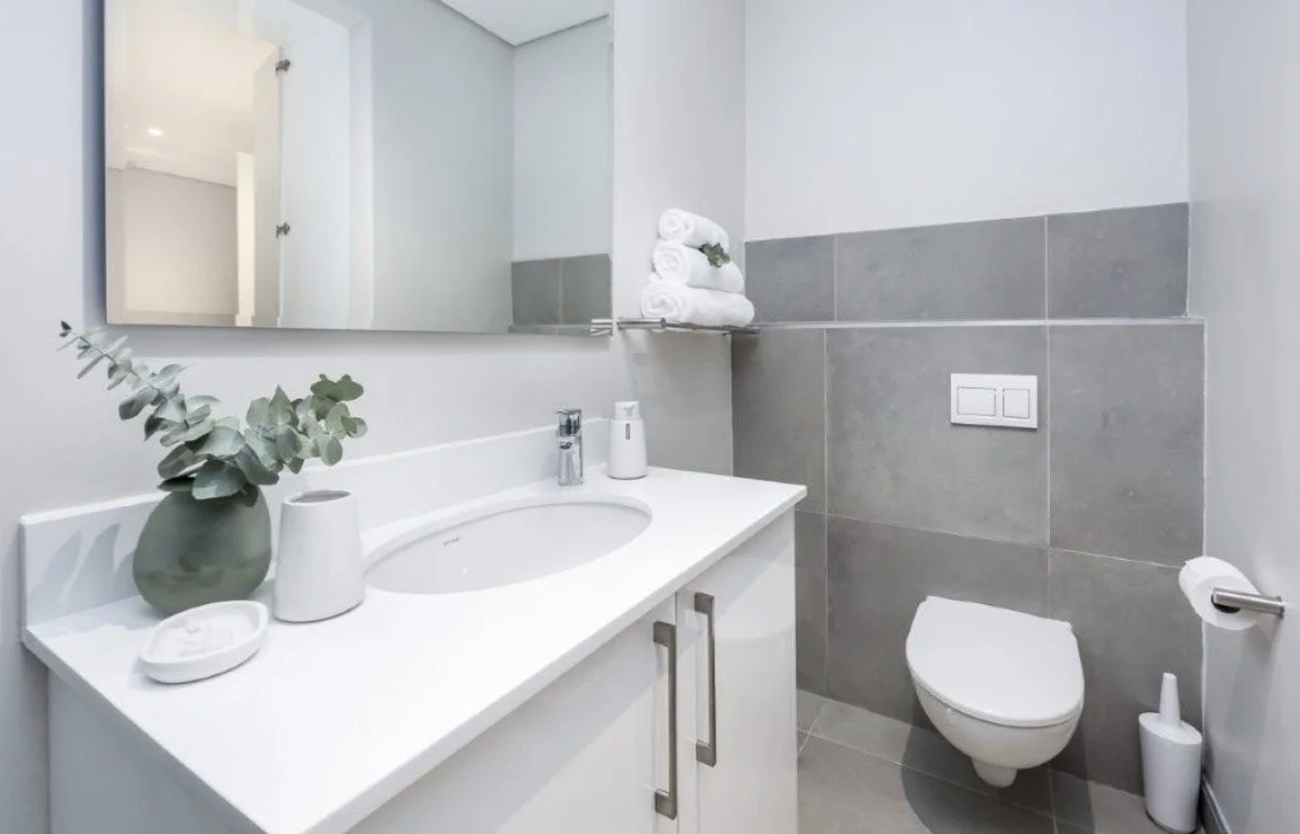 Modern bathroom with white vanity, oval mirror, gray tiled wall, underwater-style toilet, and white towels.