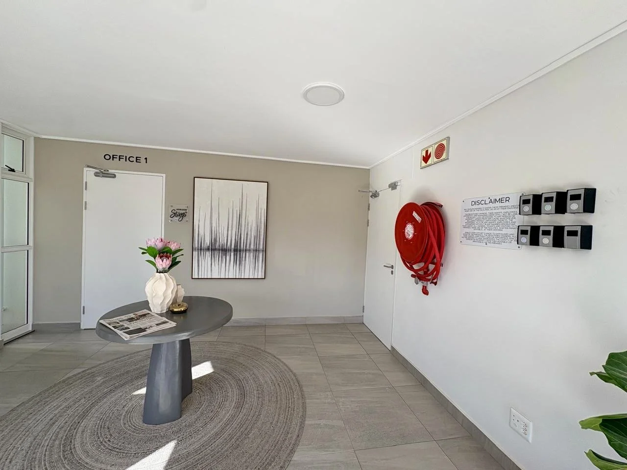Interior of a building lobby with a round table, a vase with pink flowers, a framed art piece, a fire hose reel, and warning signs on the wall.