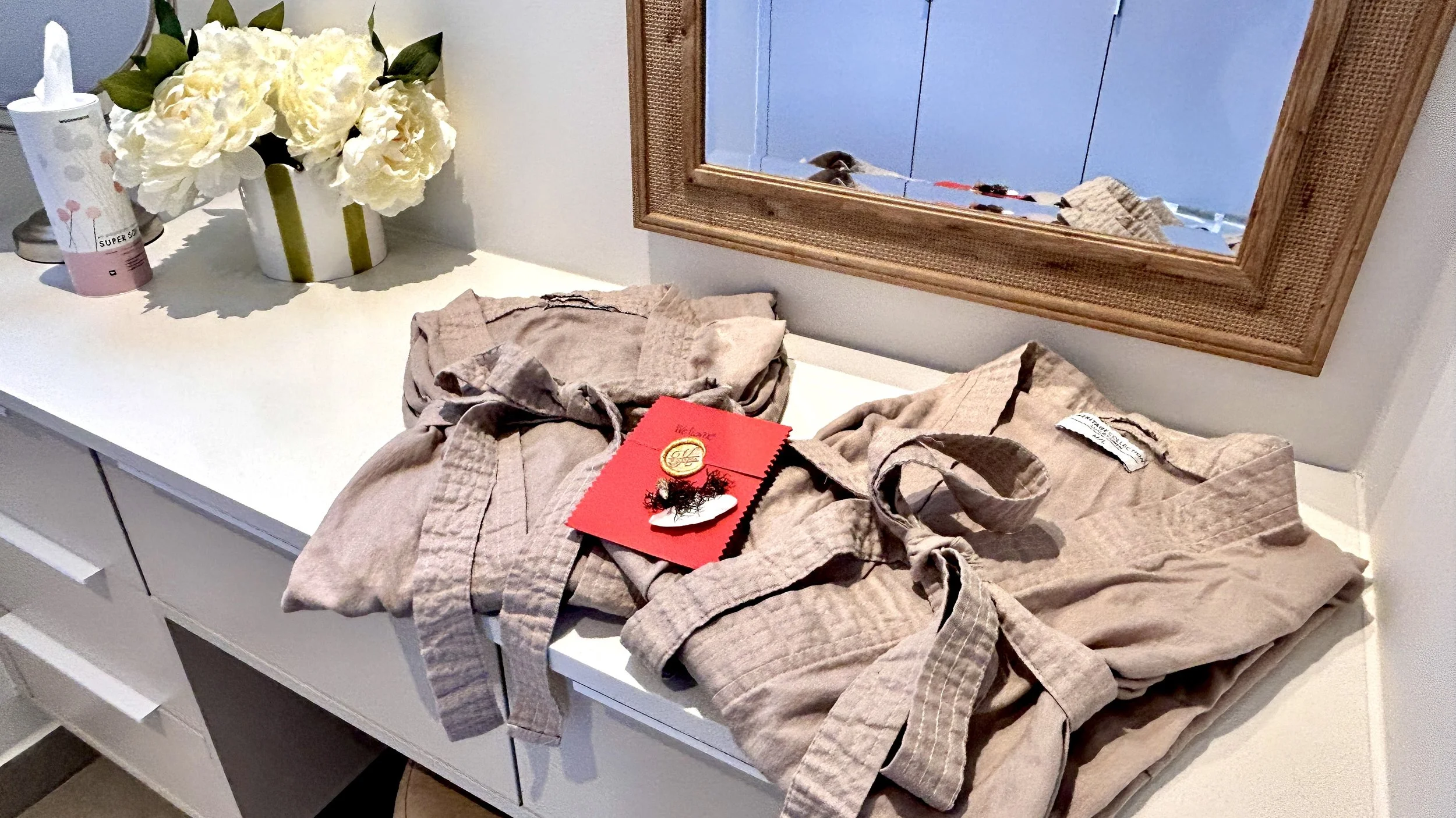 A white dresser with two beige bathrobes, a red greeting card with a gold medal pin, a pink tape dispenser, and a vase of white flowers, with a mirror reflecting part of the room.