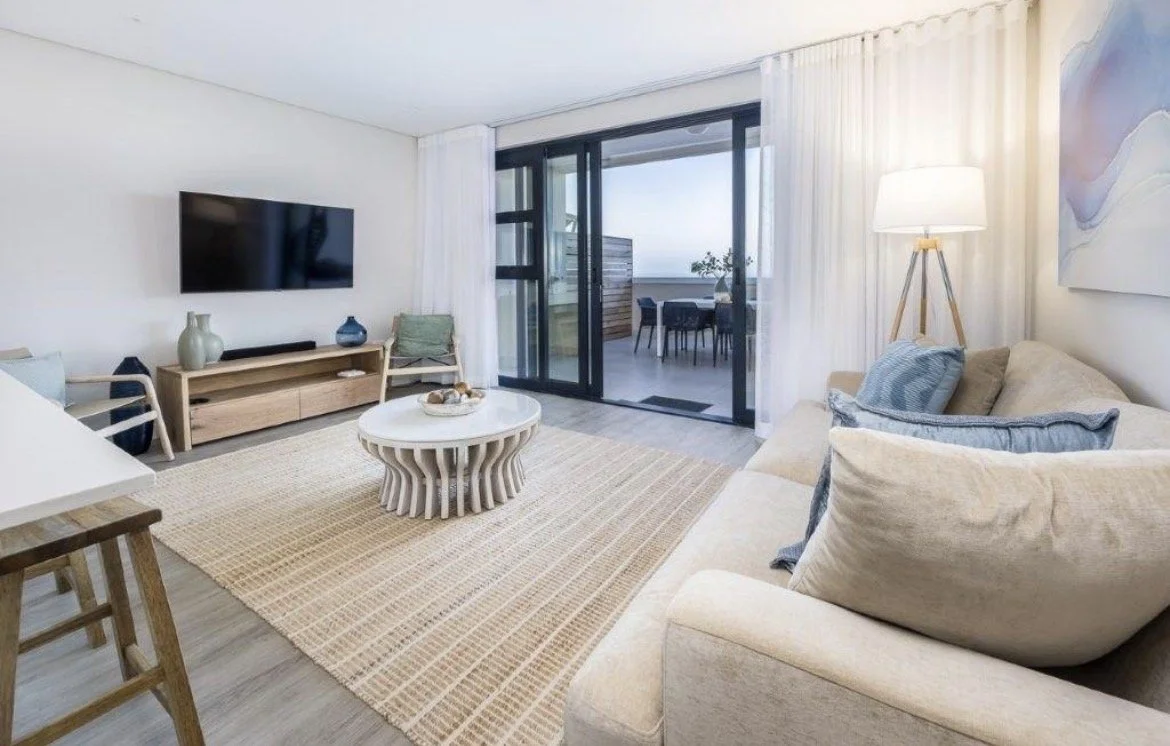 Living room with a beige sofa, a round white coffee table, wall-mounted TV, and sliding glass doors leading to a balcony with an outdoor dining area