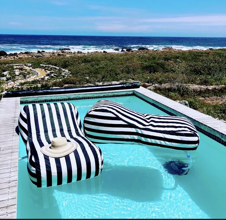 A small swimming pool with two striped pool floats, one with a hat on it, situated outdoors near the ocean with waves and green vegetation in the background.