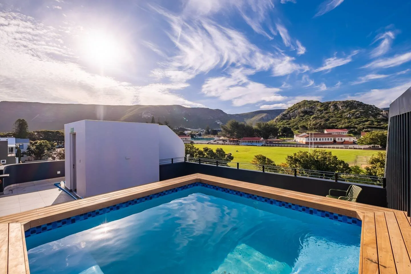 A rooftop swimming pool overlooking a grassy field, trees, and a mountainous landscape under a partly cloudy sky with the sun shining.