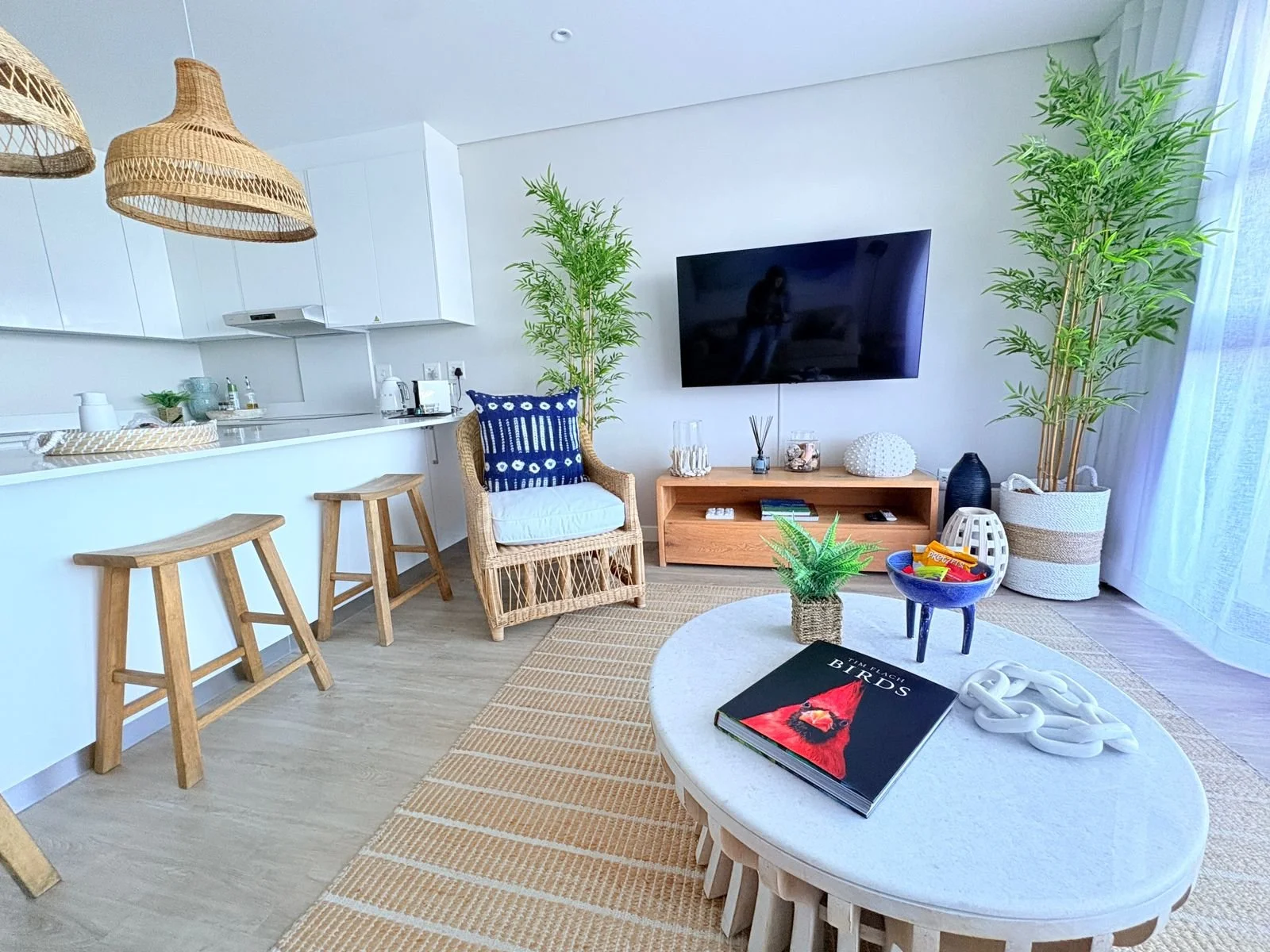 Living room and kitchen with white cabinets, wooden bar stools, wicker chair with blue pillow, wall-mounted TV, potted plants, rug, and coffee table with a magazine and decorative objects.