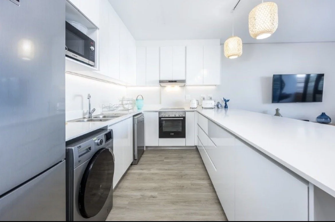 Modern white kitchen with built-in appliances, hanging pendant lights, and a large counter.