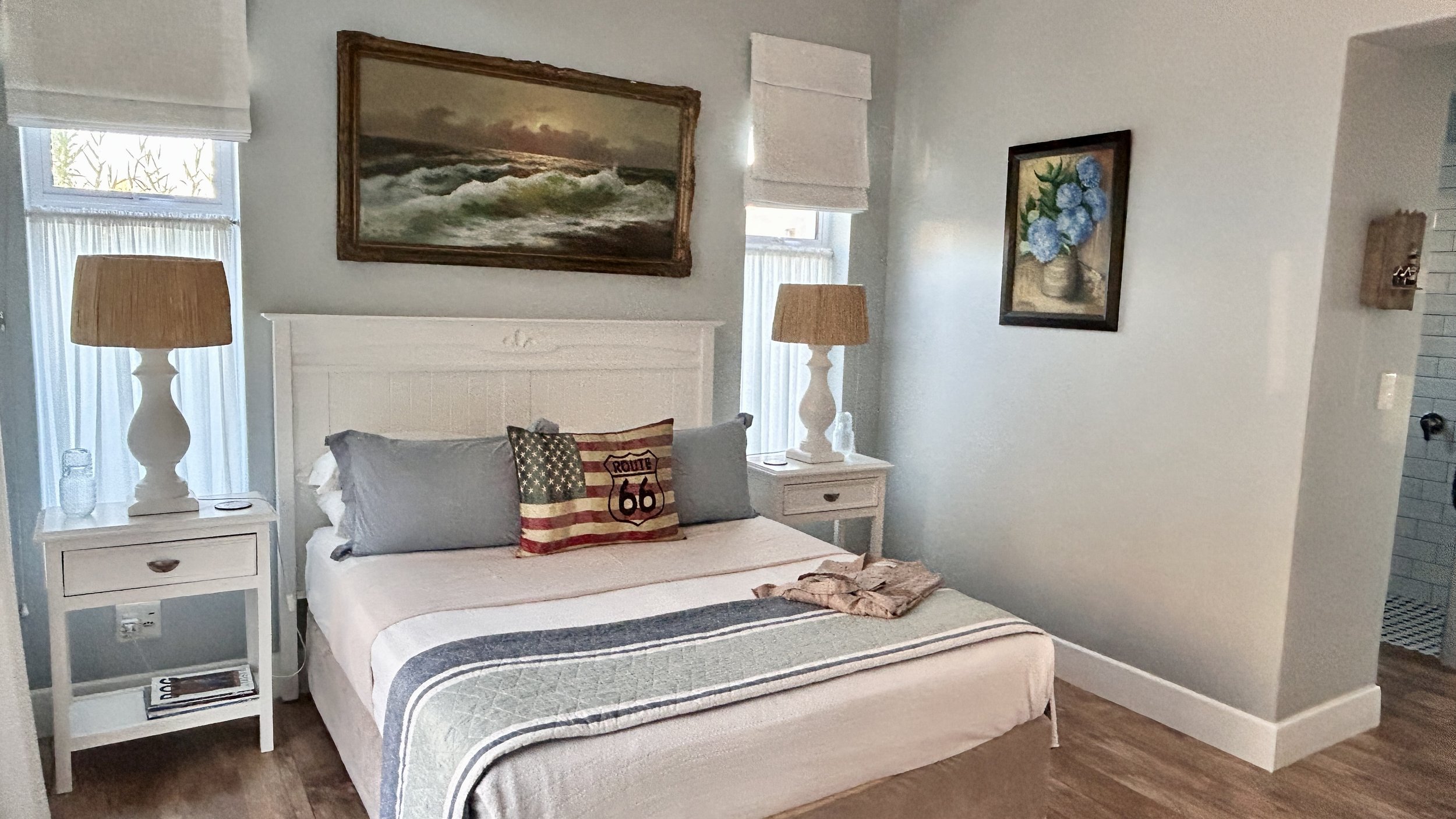A bedroom with a white bed and a throw blanket, two white nightstands with lamps, wall art including a seascape painting and a flower picture, and windows with white blinds and curtains.
