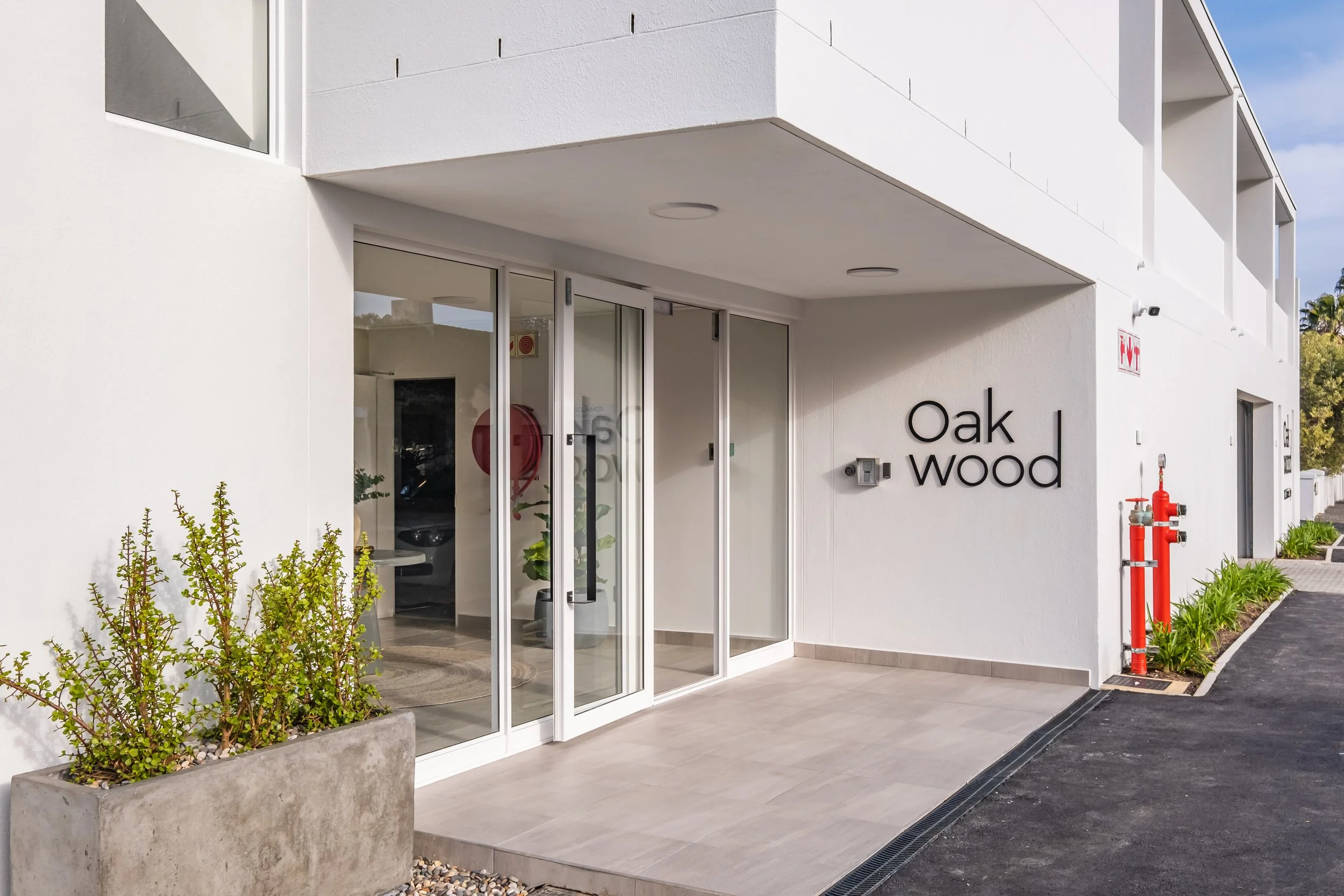 Exterior view of a modern white building with signage reading 'Oak Wood', glass entrance door, potted plants, and exterior lighting, with a parking lot and trees in the background.