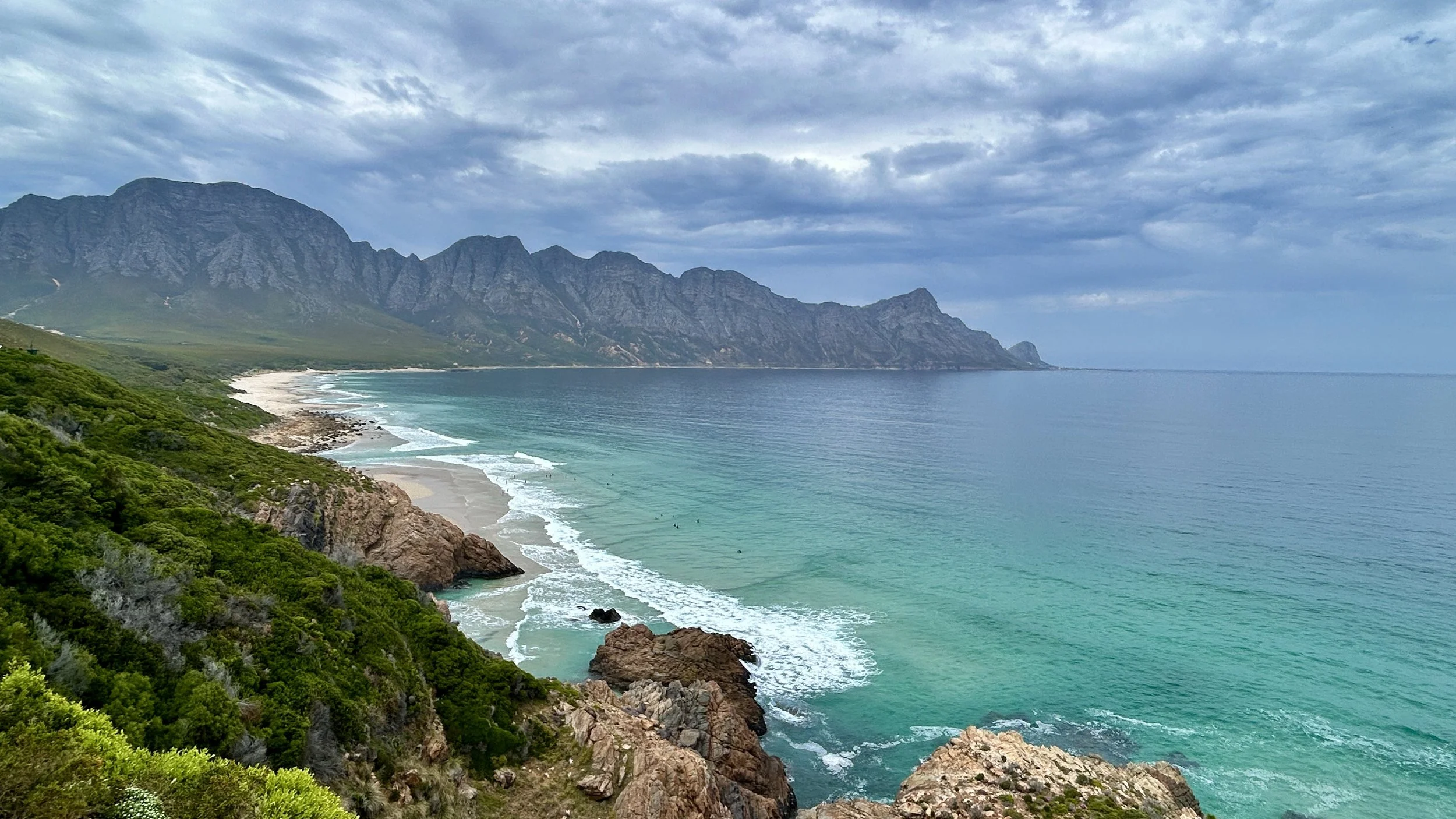 Coastal landscape with a sandy beach, rocky cliffs, green vegetation, and ocean water under a cloudy sky, with mountains in the background.