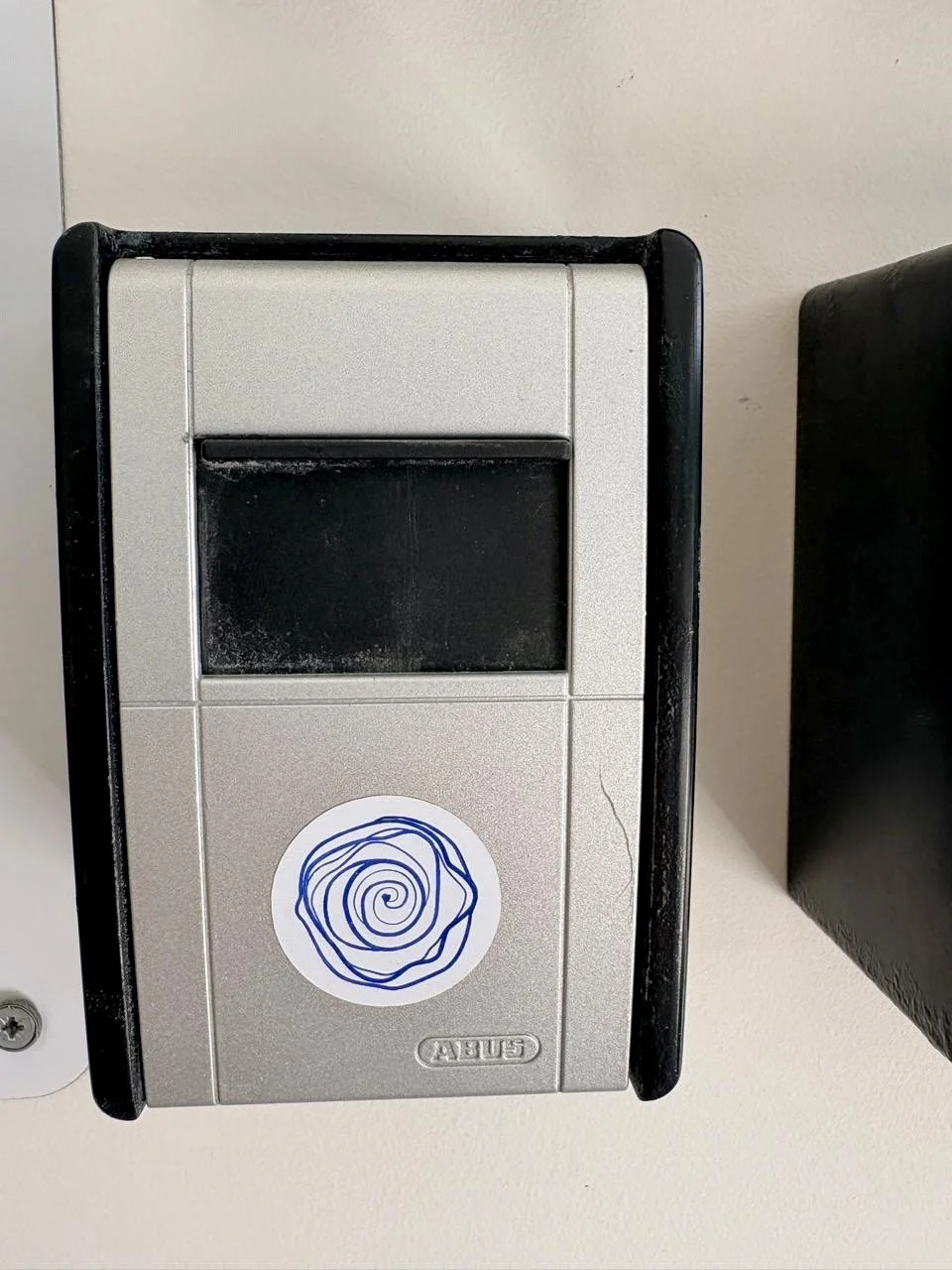 A gray and black ABUS mailbox with a circular blue abstract sticker on the front. The mailbox is mounted on a white wall.