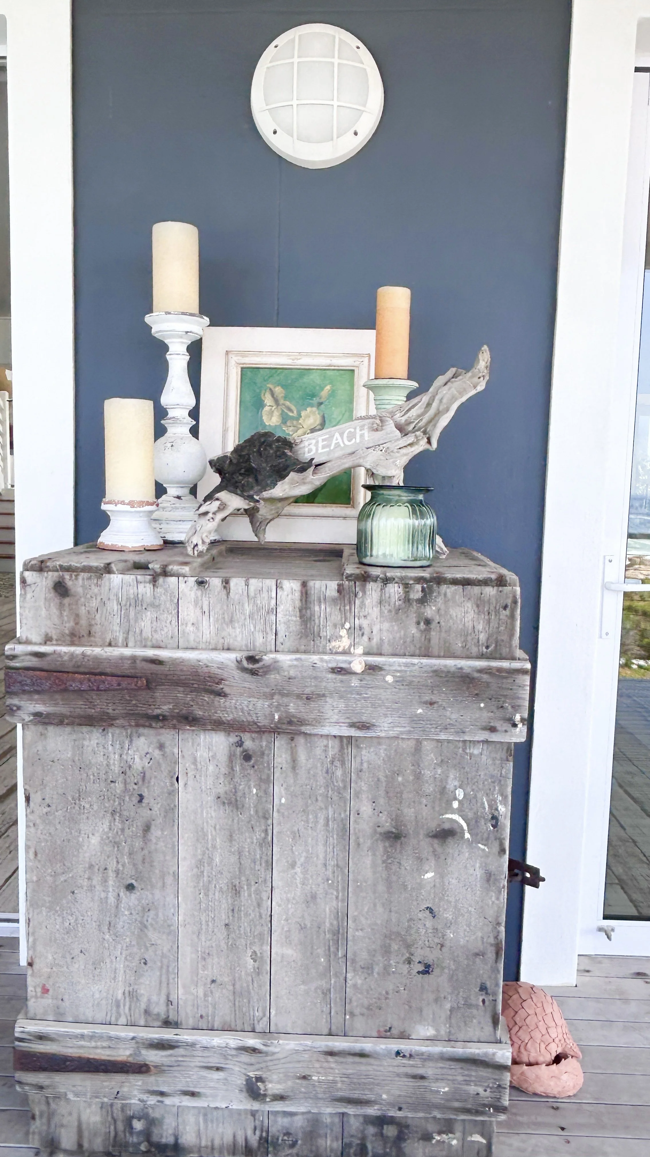 Decorative porch display with candles, framed butterfly painting, driftwood with 'BEACH' sign, and weathered wooden storage box.