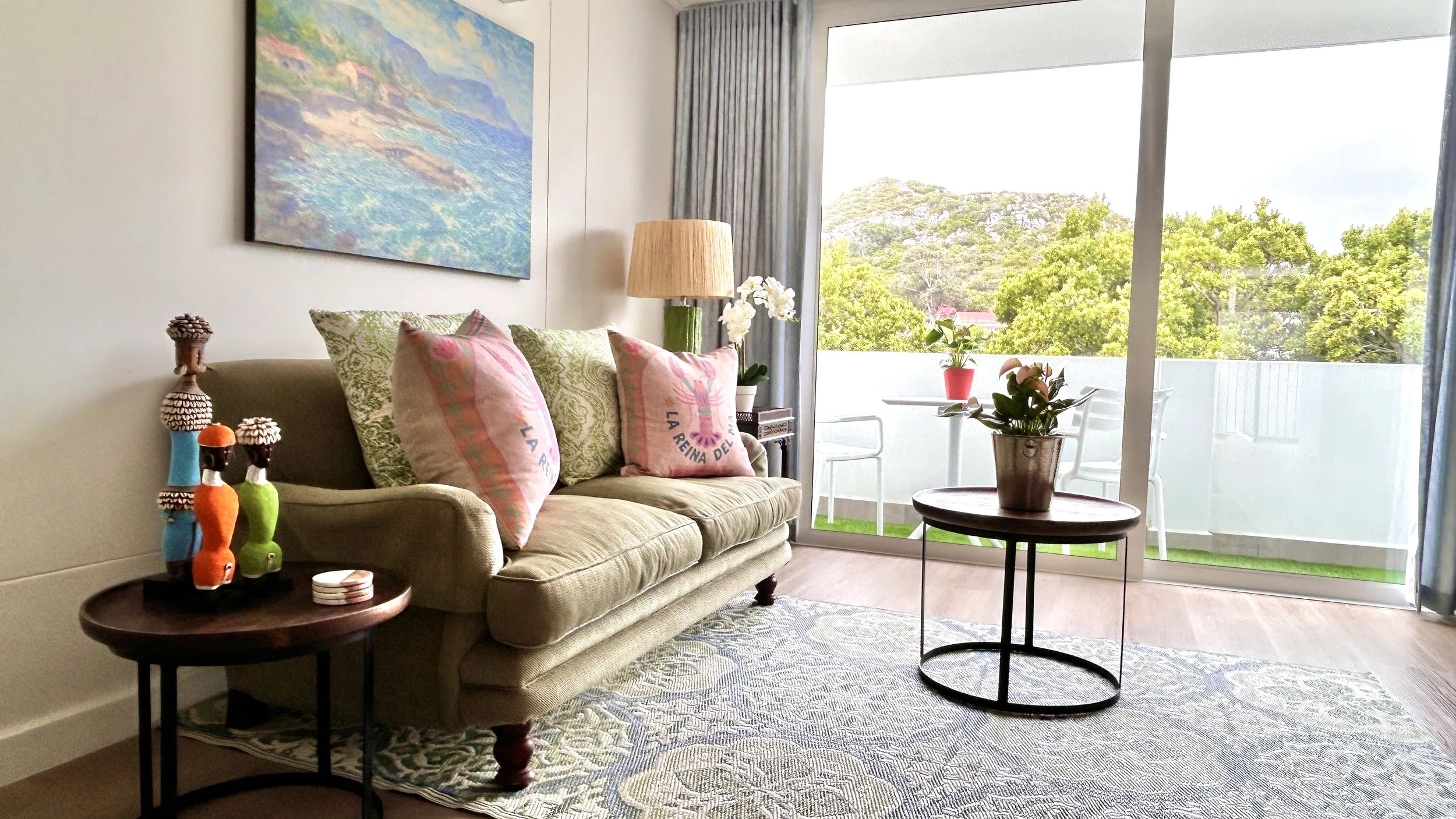Living room with beige sofa, pink cushions, a side table with decorative bottles, a lamp, a wall art, and a balcony with potted plants and greenery outside.