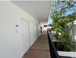 Exterior corridor of an apartment building with white walls and doors, numbered 102, brown wooden flooring, black railing, and green trees outside.