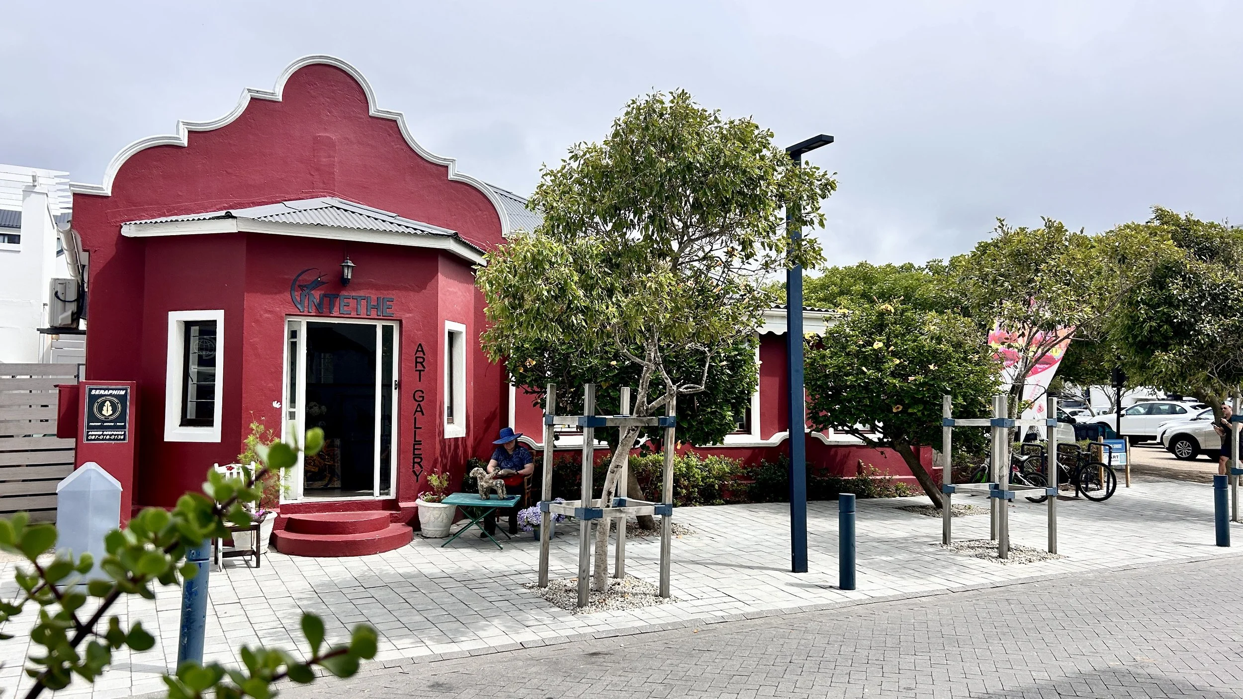 A red art gallery building named 'INTETHE' on a street with trees, a person sitting on a bench, bicycles, and parked cars in the background.
