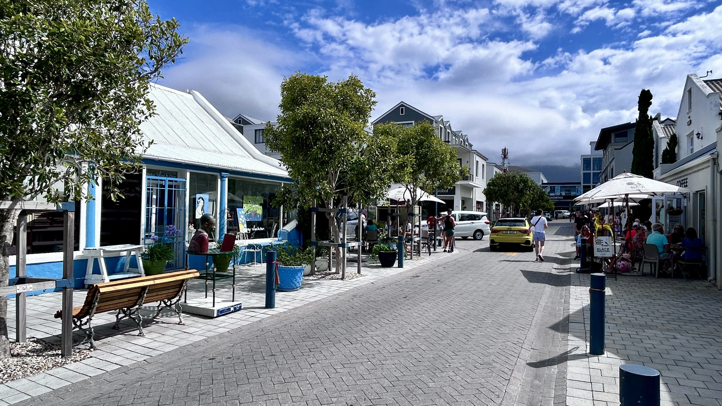 A lively outdoor shopping street with shops, cafes, and people walking, some sitting under umbrellas, trees lining the sidewalk, and a bright yellow car driving on the paved street. The sky is partly cloudy with blue patches.